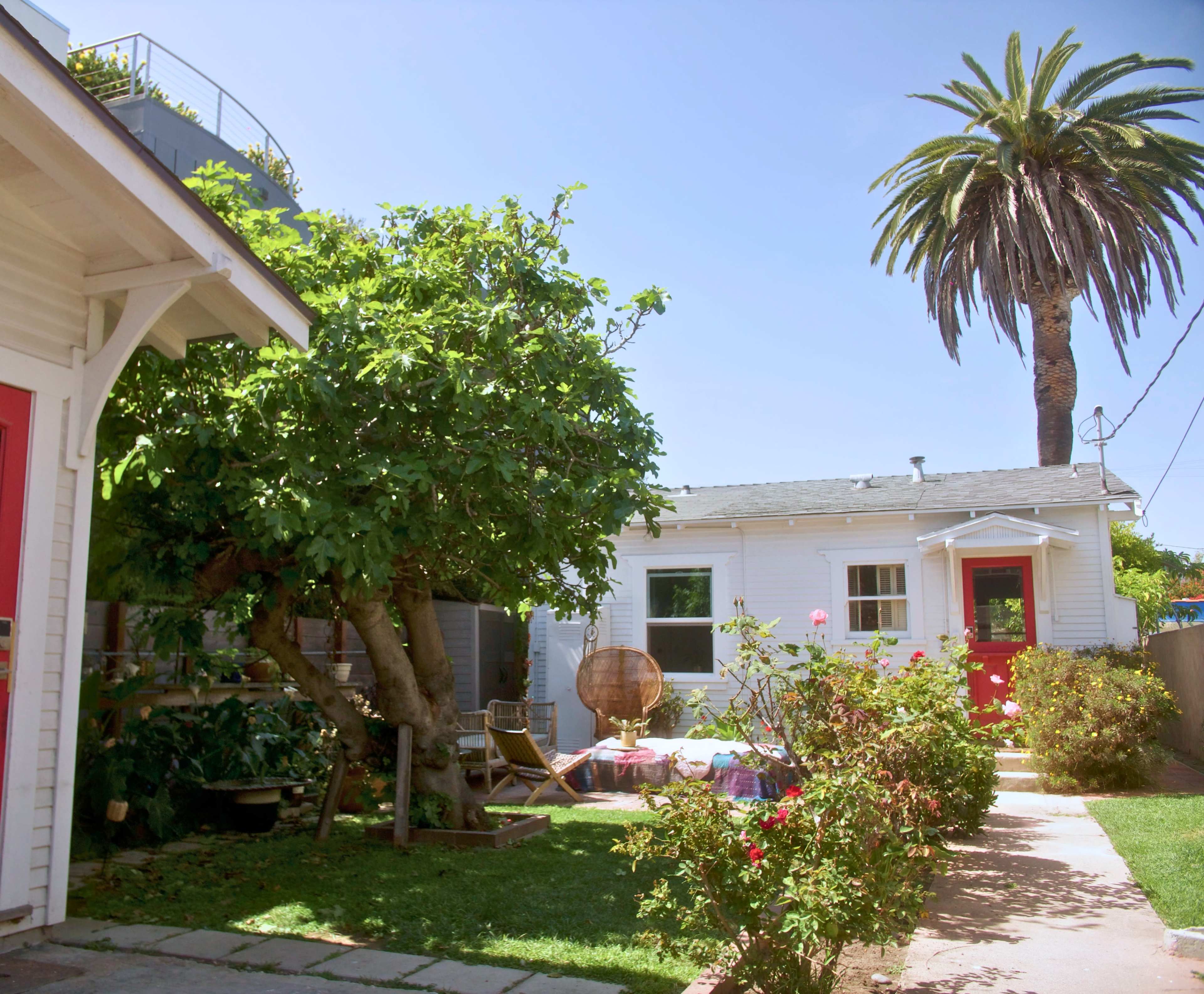 The image shows a small, white house with a red door, surrounded by green trees and a well-maintained garden in a sunny setting.