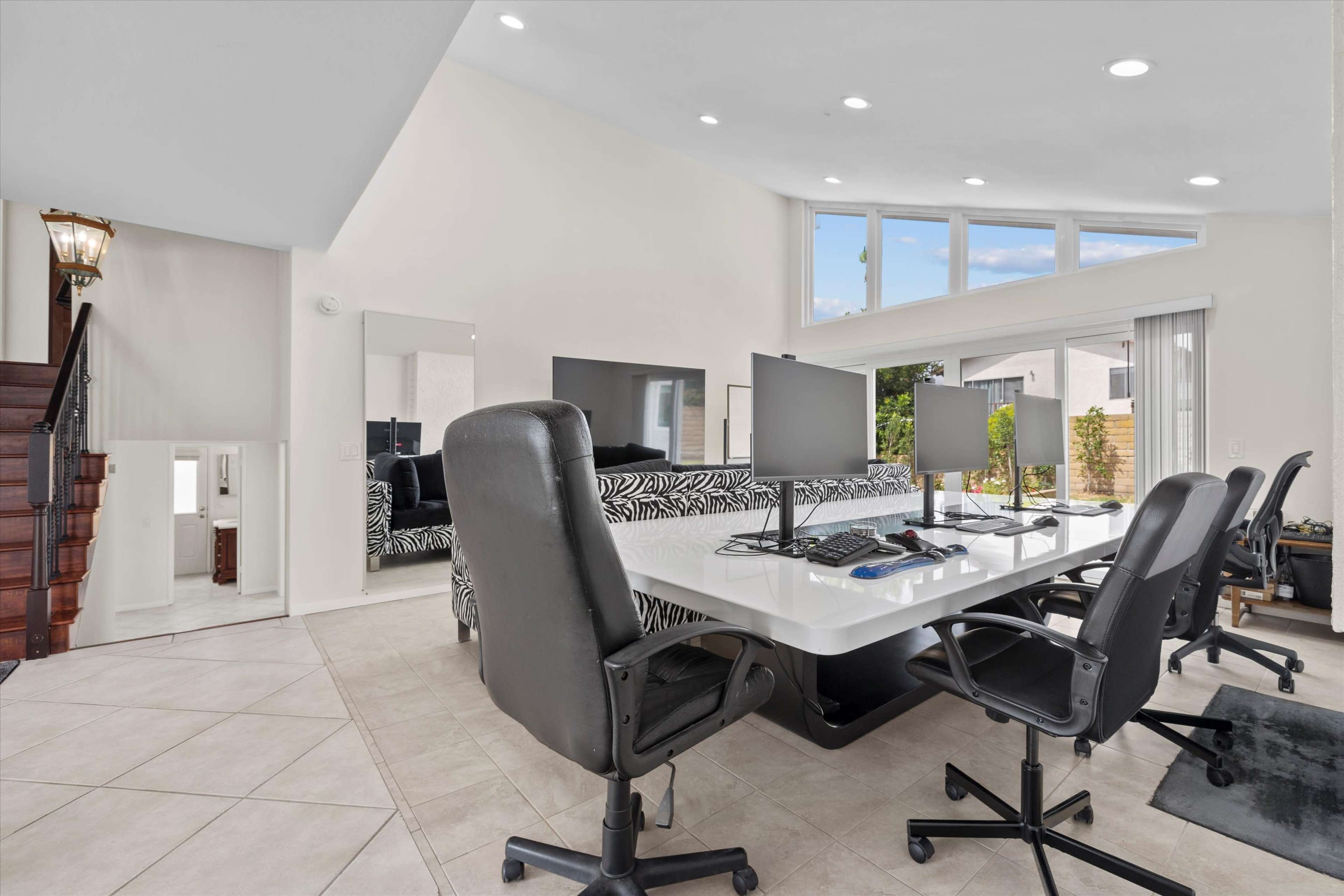 The image shows a modern, open-concept home office space with multiple computer workstations, sleek furniture, and large windows allowing natural light.