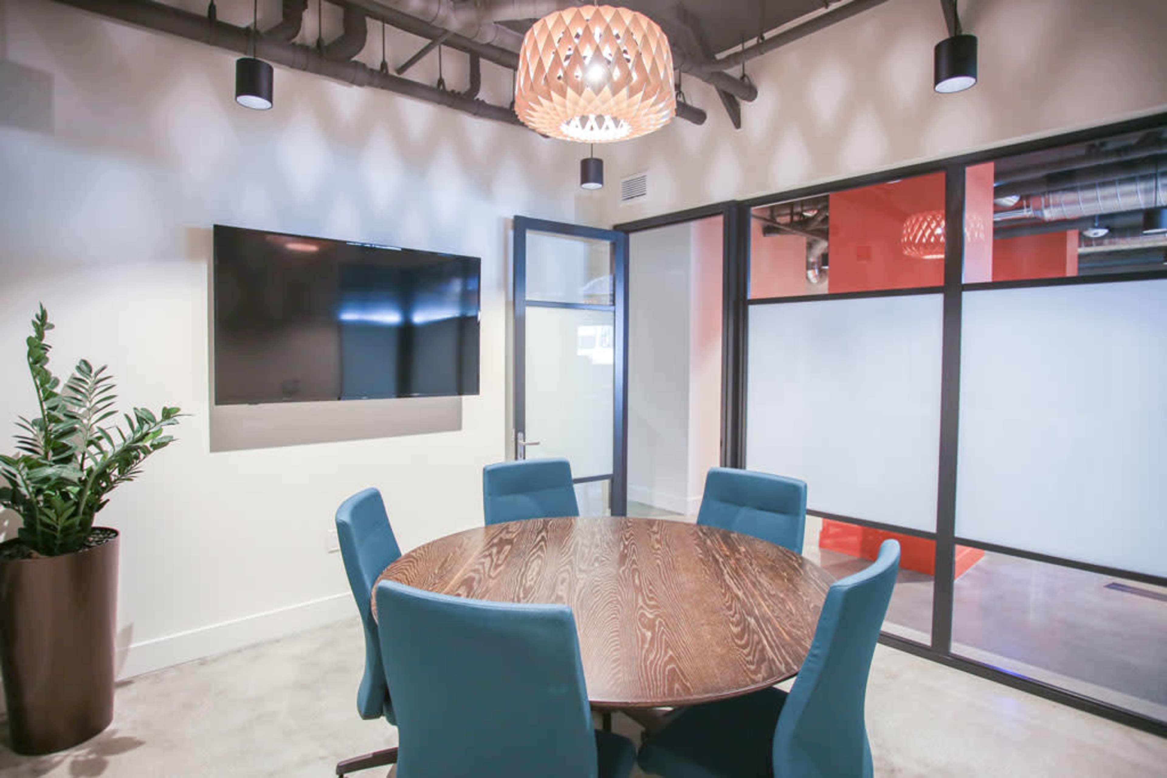 A conference room features a round wooden table surrounded by blue chairs, with a television mounted on the wall and glass partitions.