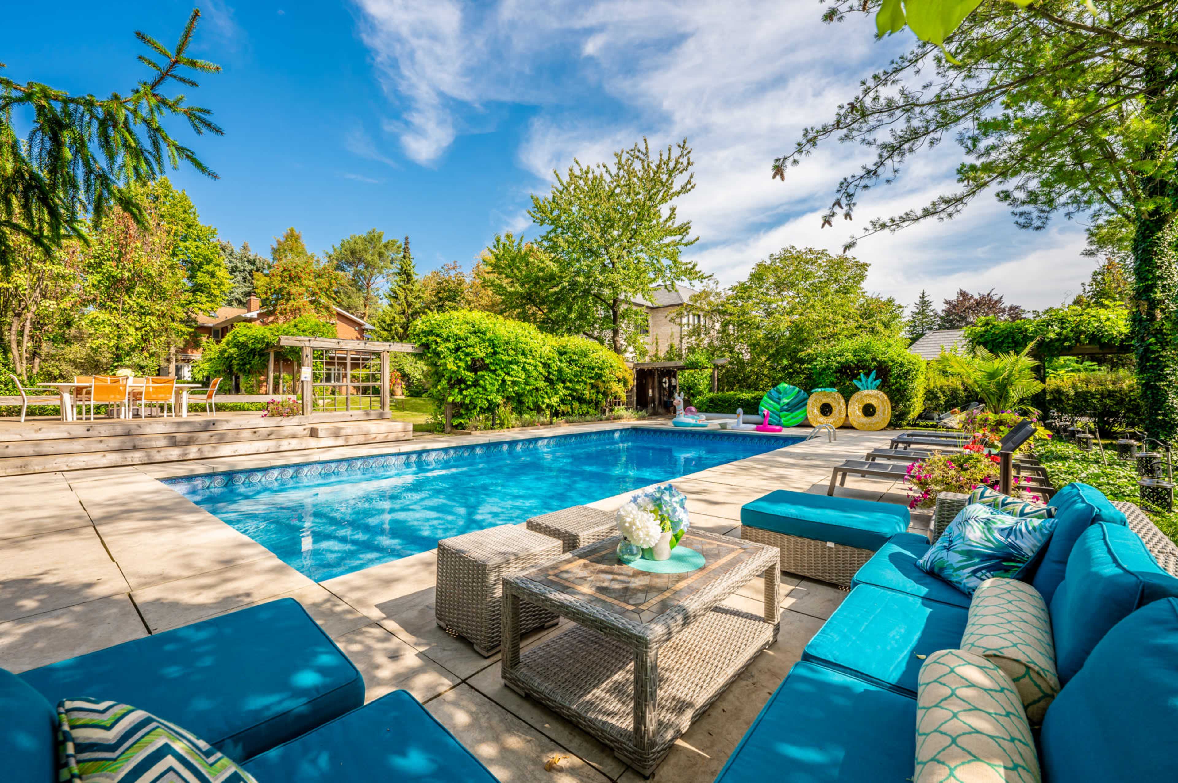 A backyard swimming pool surrounded by lounge seating and greenery, with a patio and dining area visible in the background.