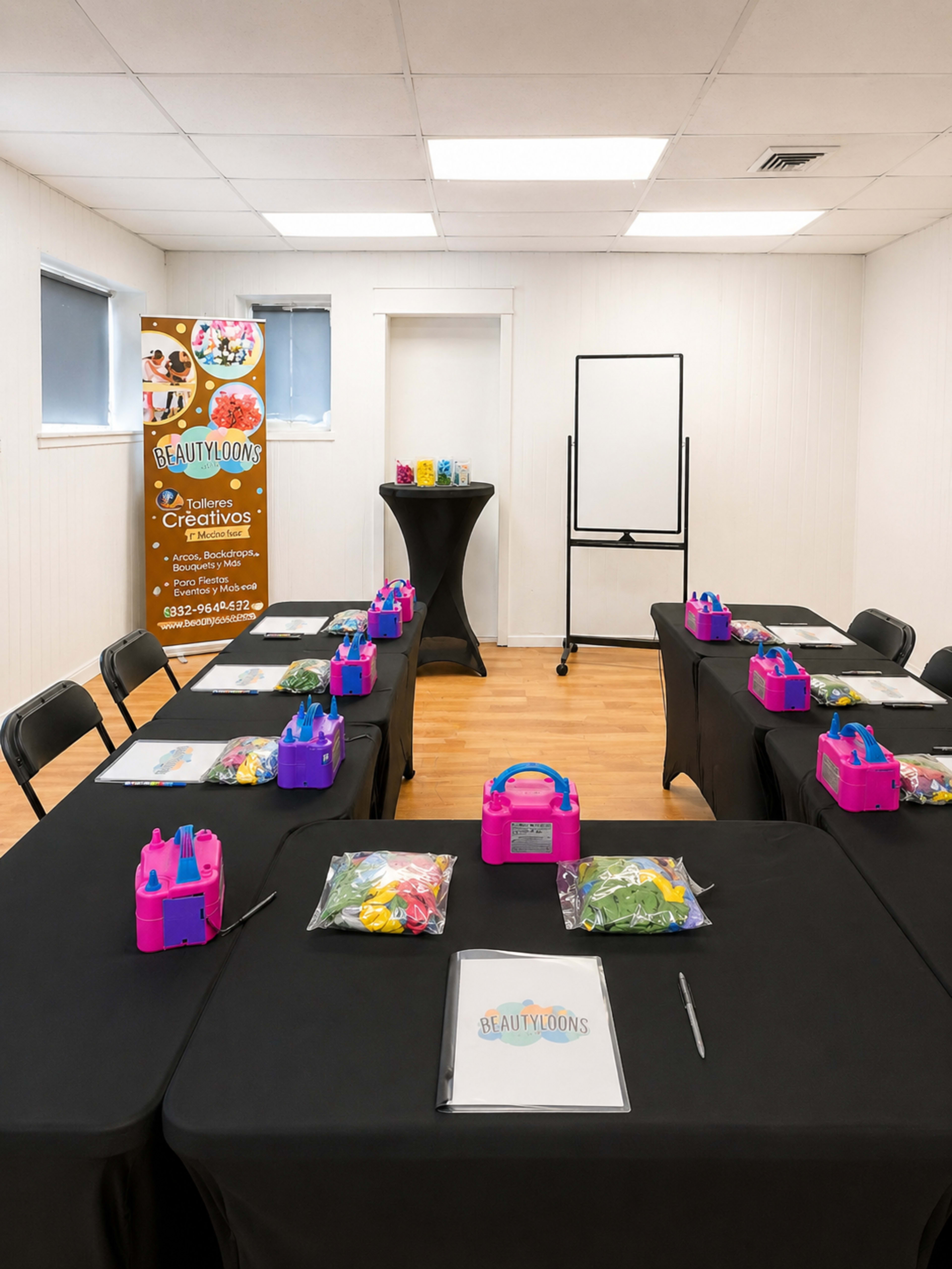 The image shows a spacious room set up with multiple tables, each covered in black tablecloths, arranged with colorful kits and supplies for a crafting or decoration activity, with a banner for "Beautyloons" in the background.