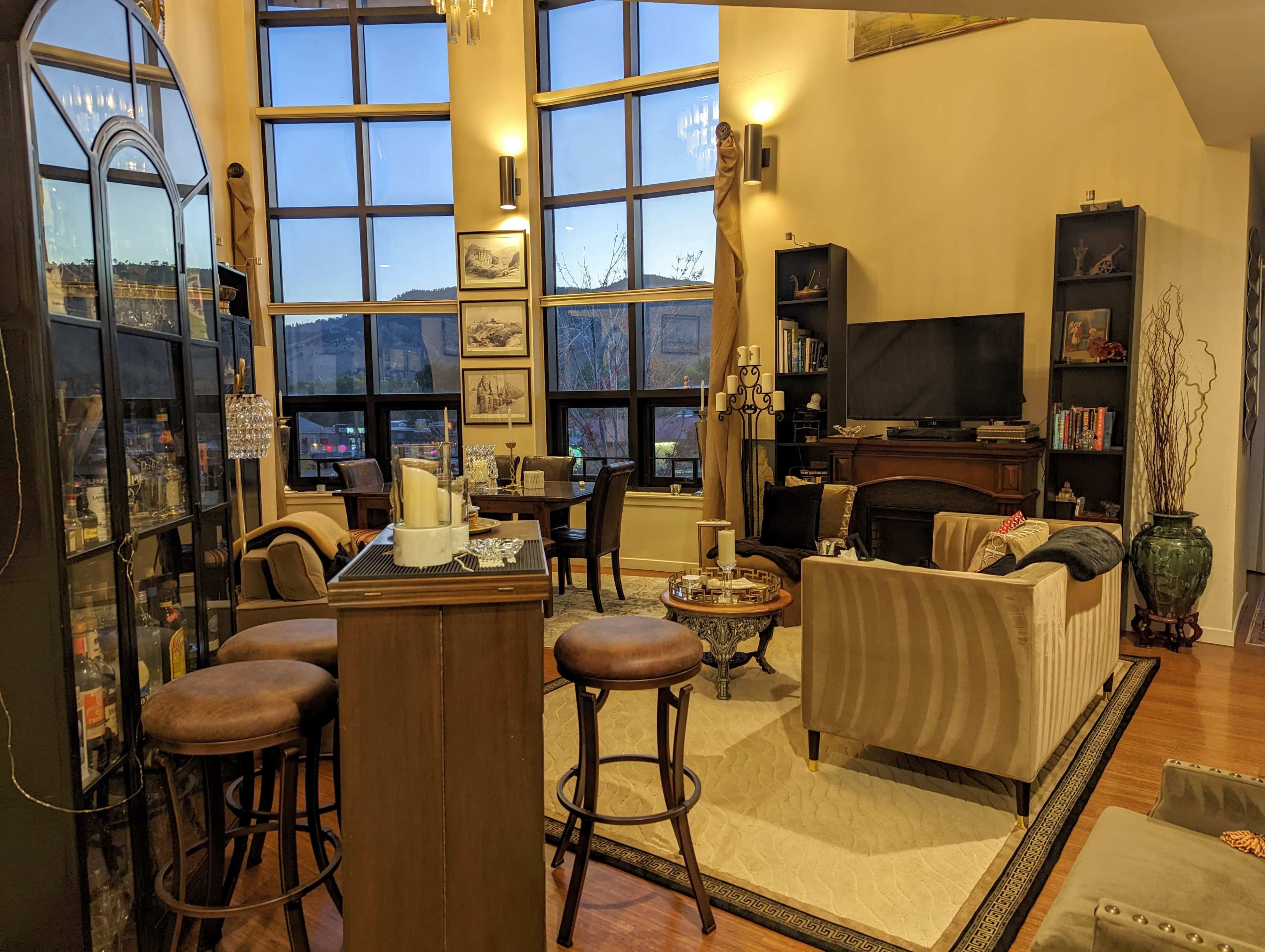 The image shows a modern living room with large windows, a seating area featuring a sofa and chairs, a bar with stools, and various decorative items on shelves.