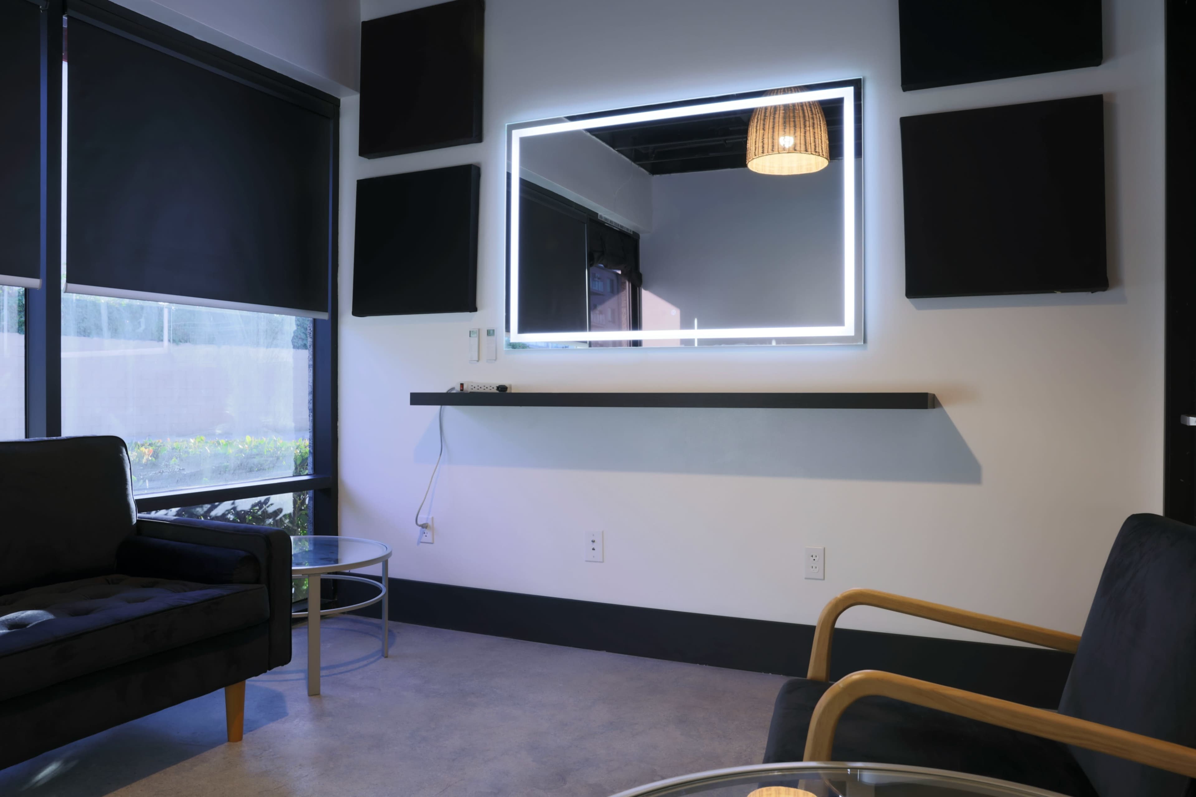 A minimalistic room features a mirror with a built-in light above a shelf, flanked by two black chairs and a glass table.