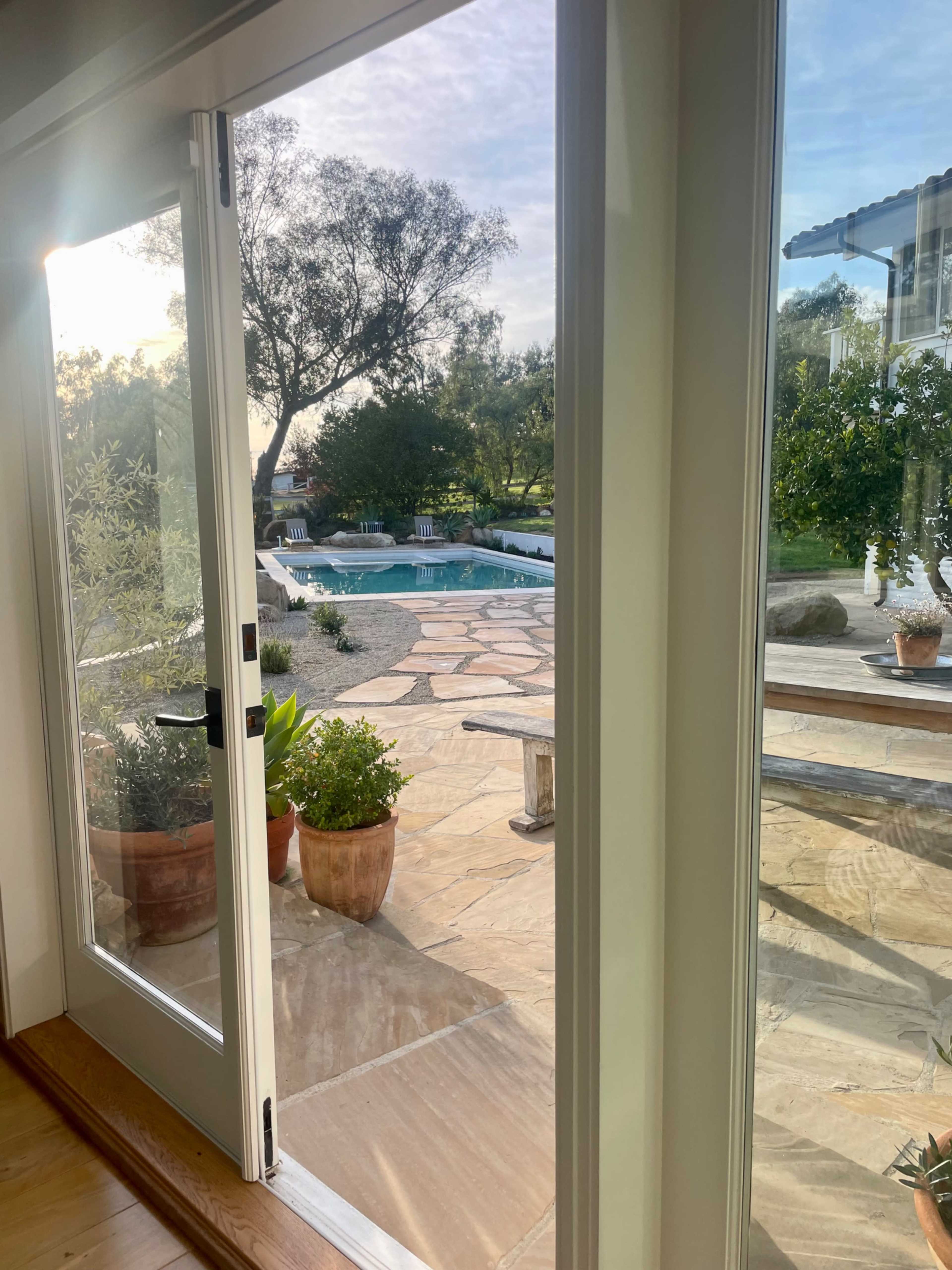 The image shows a view of a pool and patio area through an open glass door, surrounded by trees and gardens.