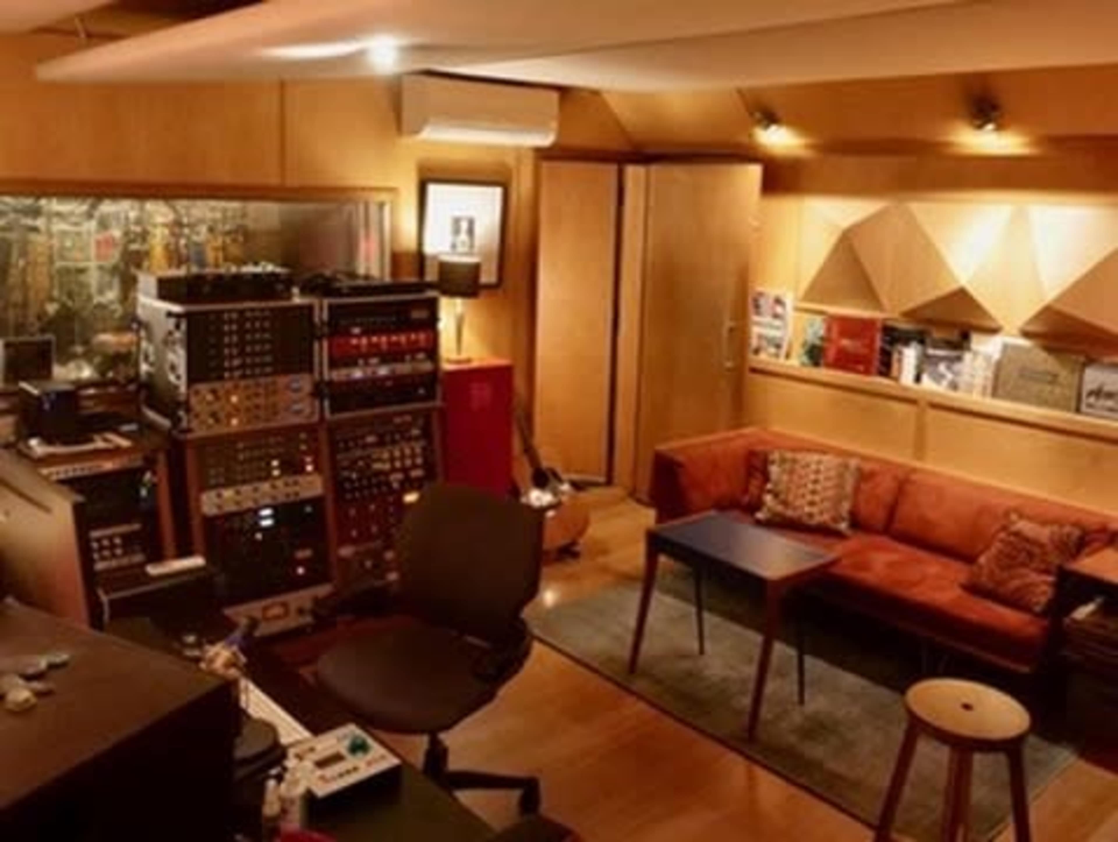 The image shows a sound recording studio with a cozy seating area, various audio equipment, and soundproofed walls.