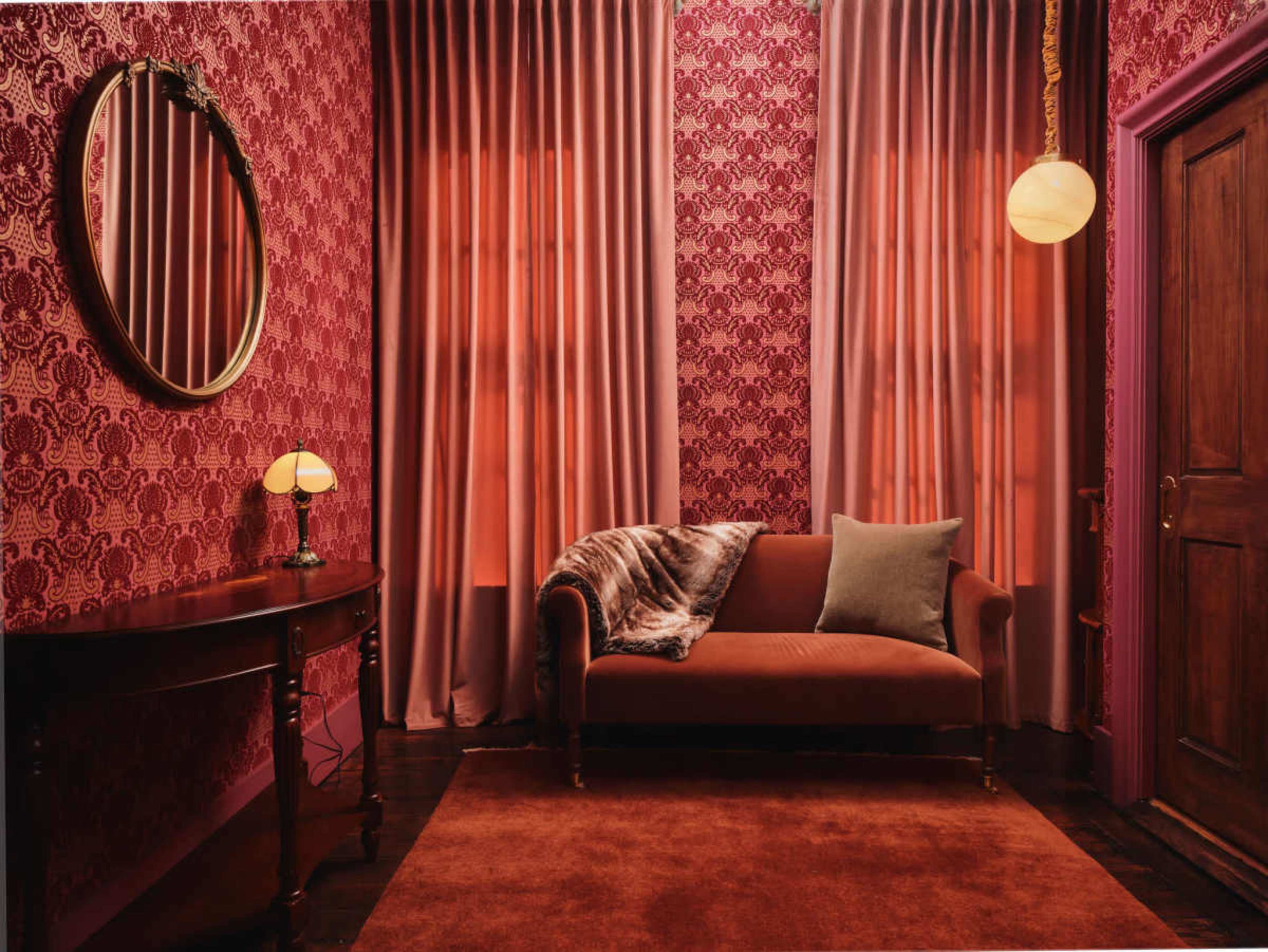 The image depicts a dimly lit room with red patterned wallpaper, featuring a brown sofa, a small lamp, and a round mirror on the wall.