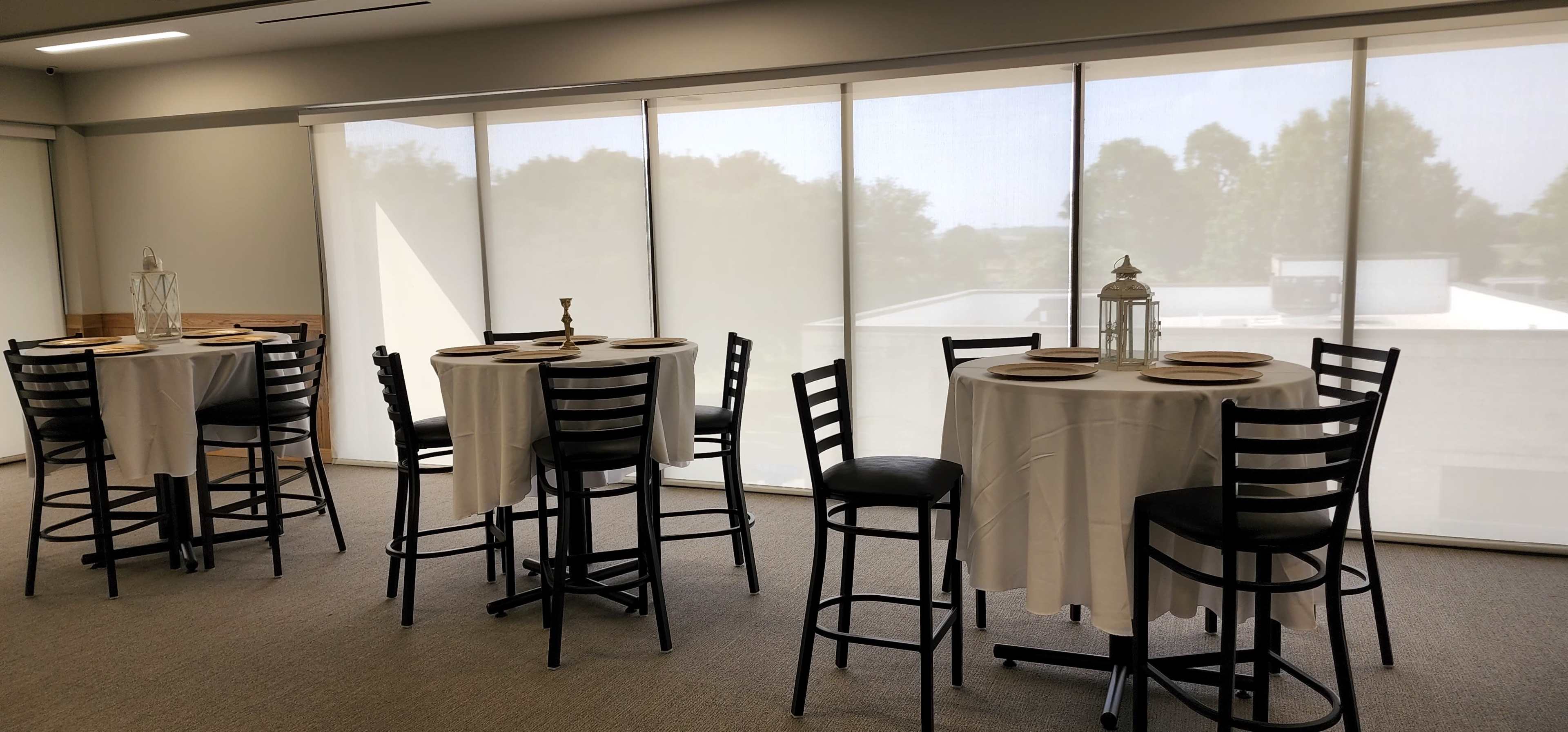 A room with several round tables covered in white tablecloths, set with plates, and surrounded by black chairs, with large windows allowing natural light to enter.