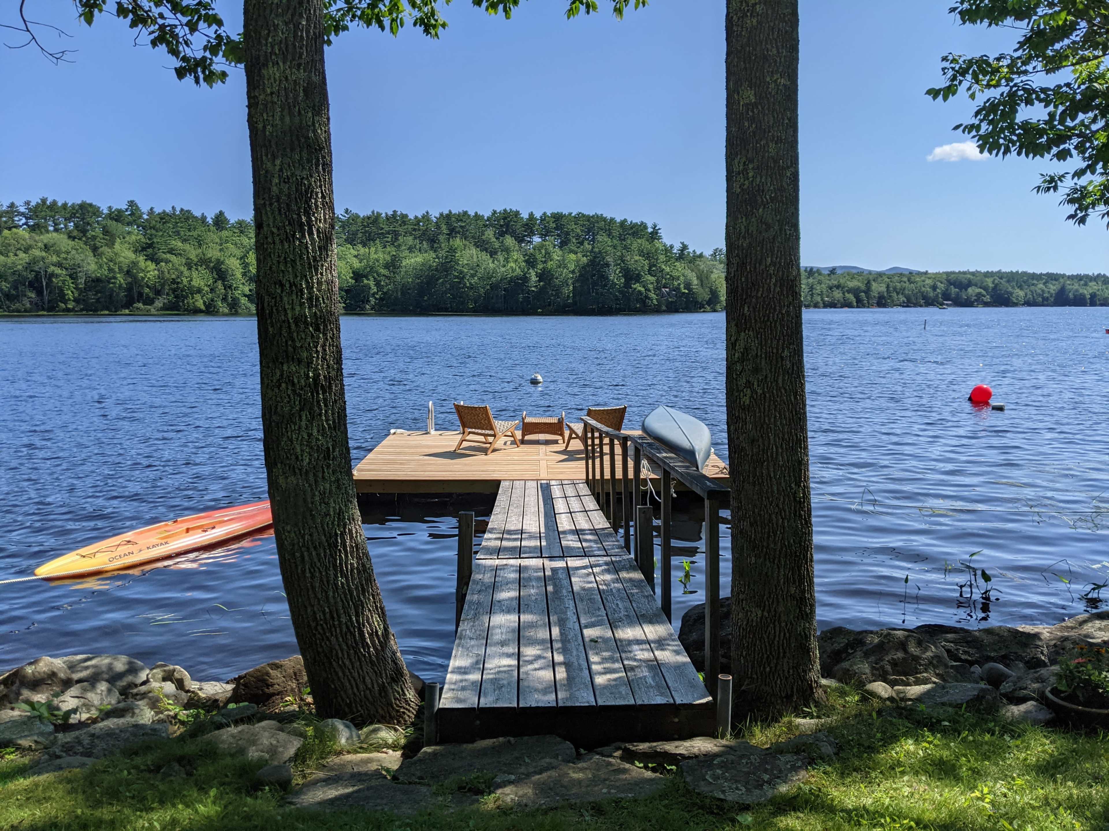 A wooden dock extends into a lake, flanked by two trees, with chairs and a kayak resting nearby.