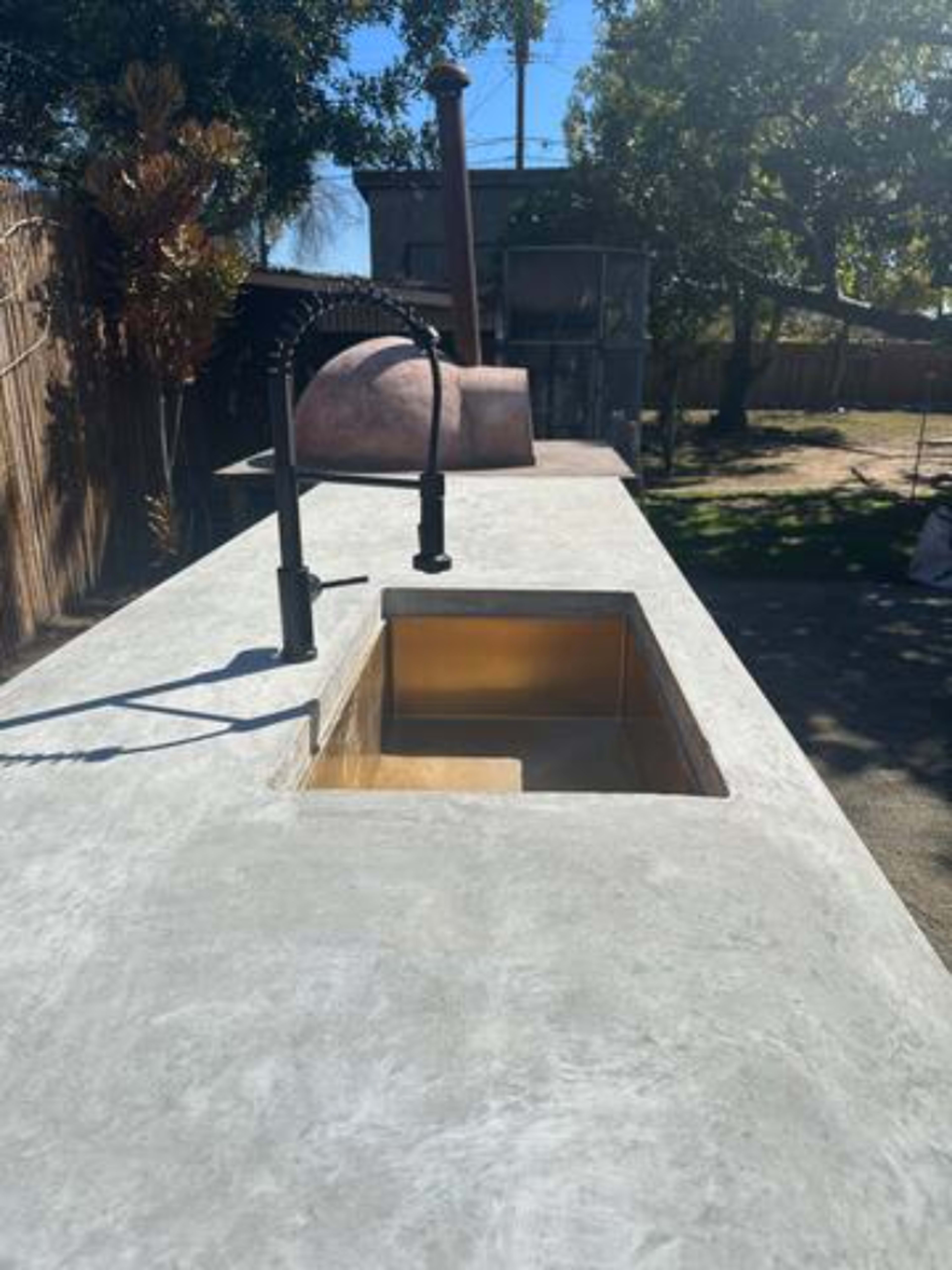 The image shows a concrete outdoor countertop with a sink and faucet, set in a backyard with a large tree and a building in the background.