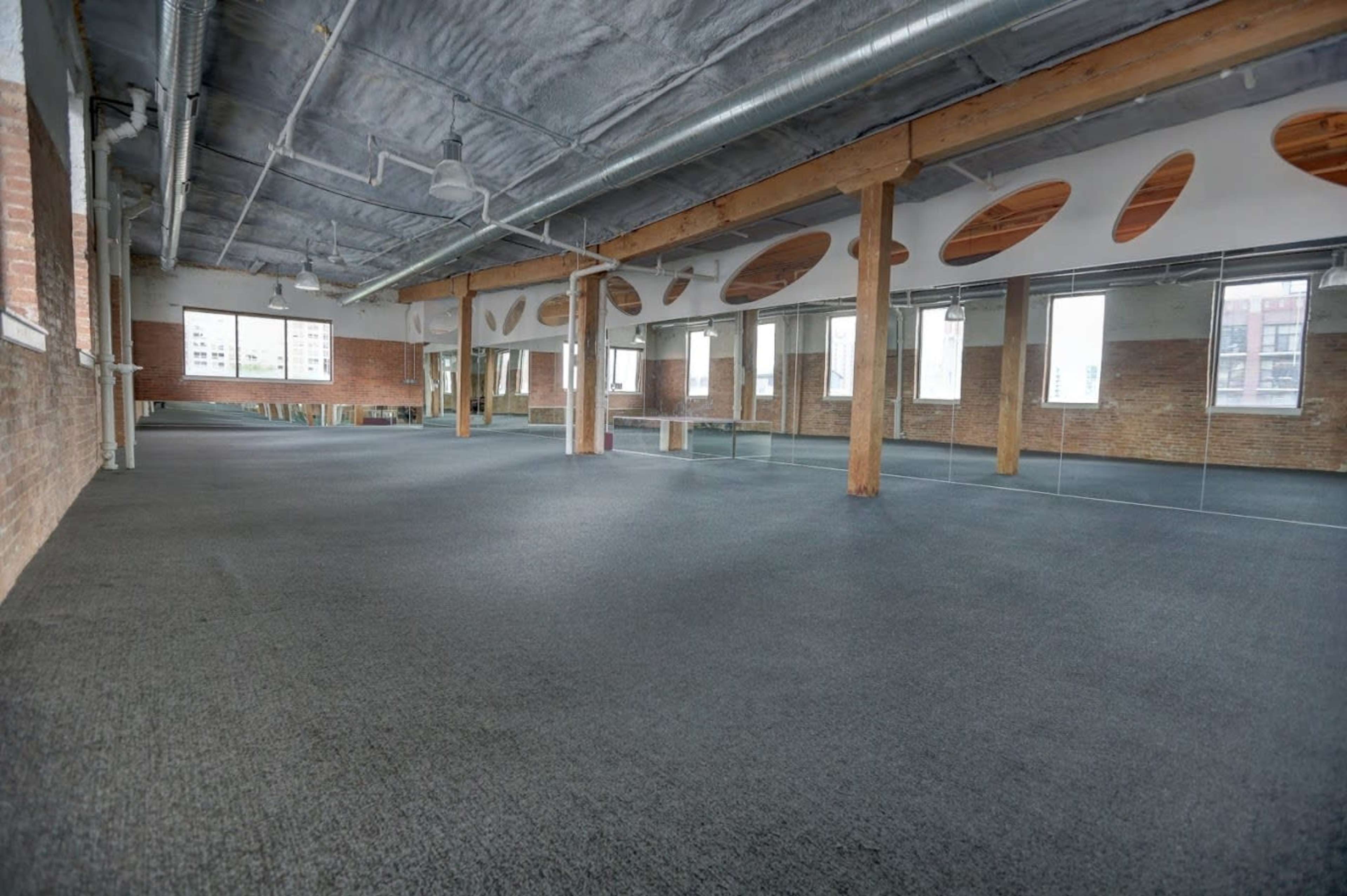 A large, empty room with wooden columns and mirror-covered walls, featuring gray carpet and multiple windows.