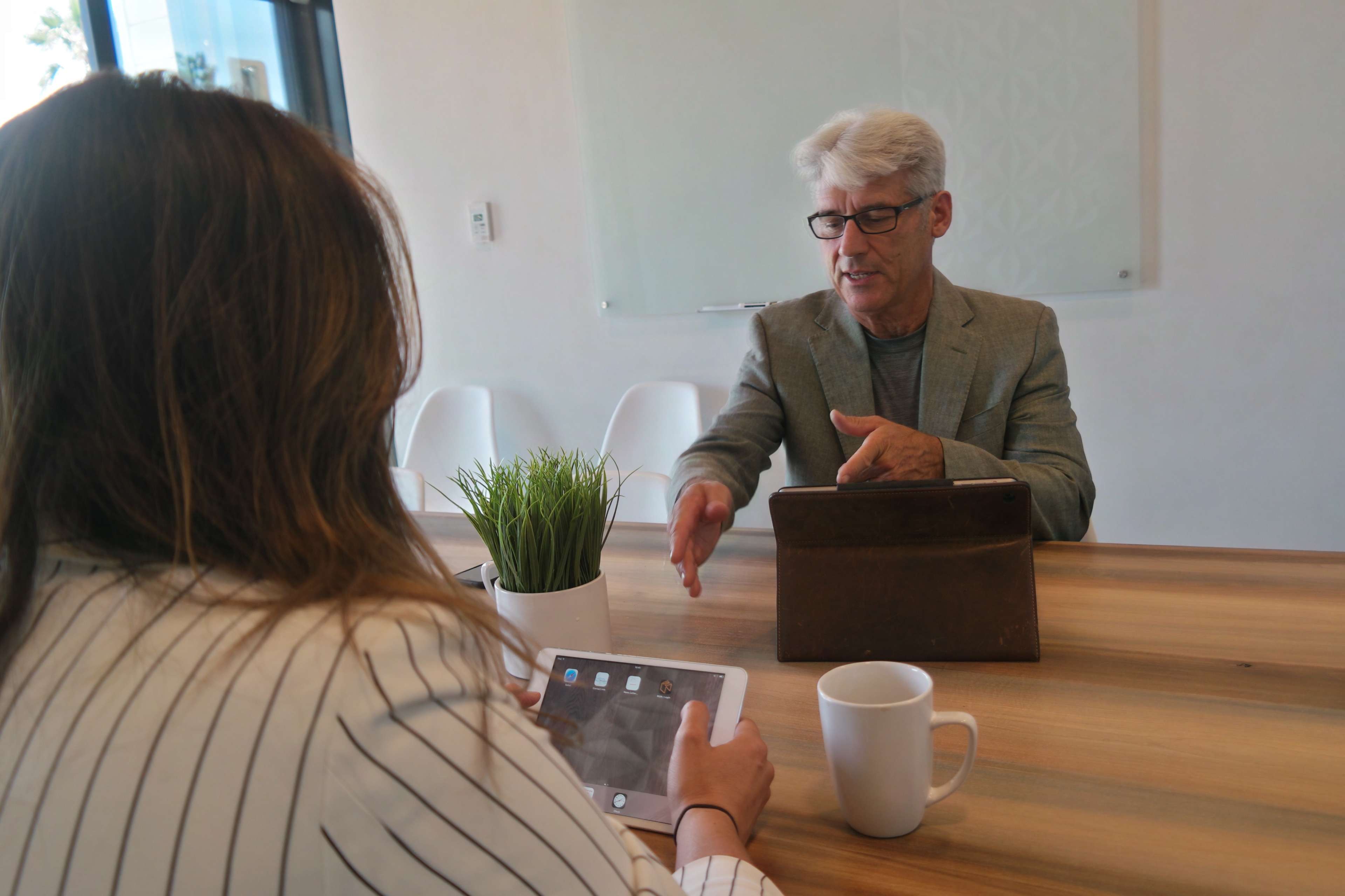A man in a gray blazer gestures while speaking to a woman sitting at a wooden table, holding an iPad, with a coffee mug nearby.