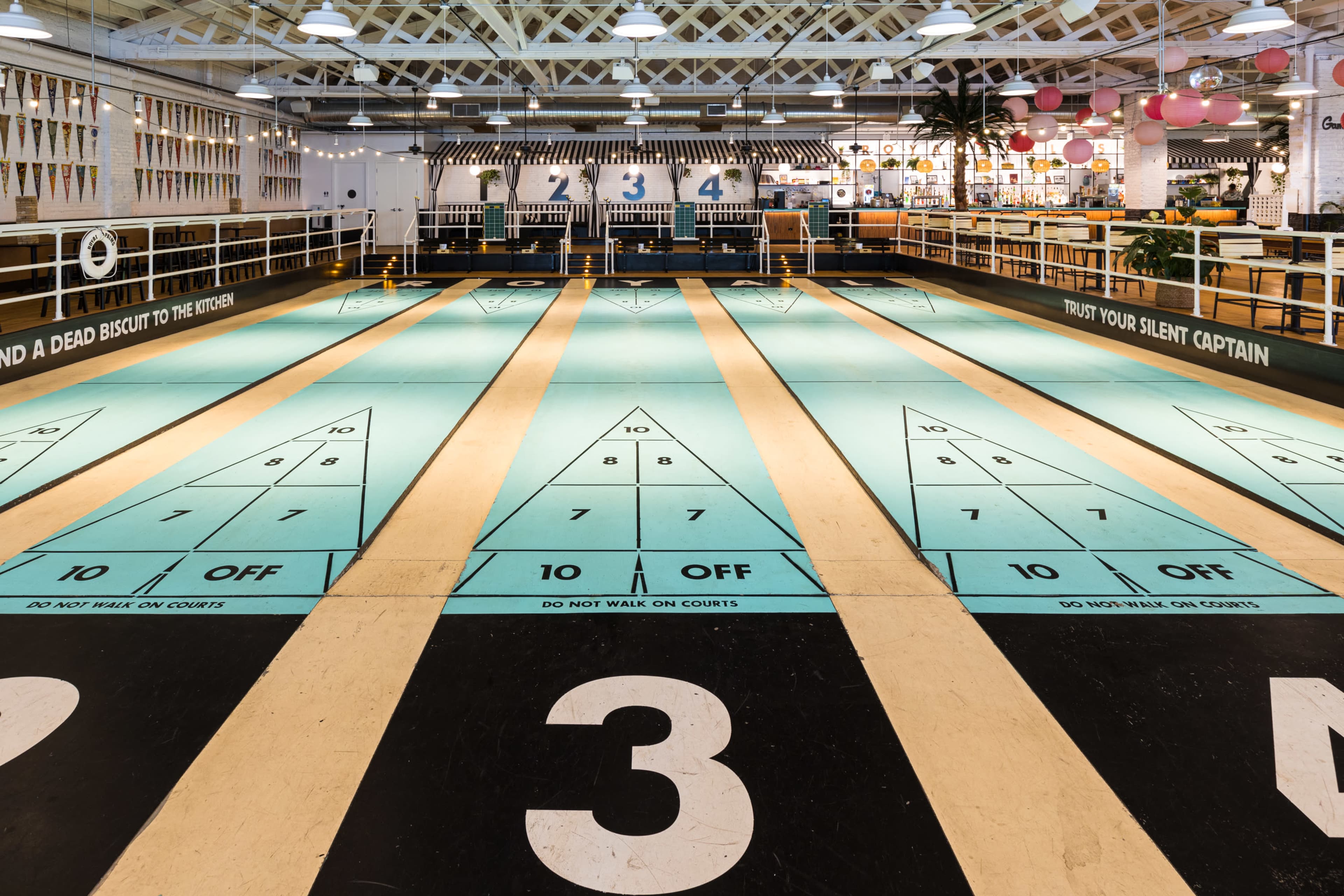 The image shows a brightly lit indoor shuffleboard court with numbered lanes and decorative signage.