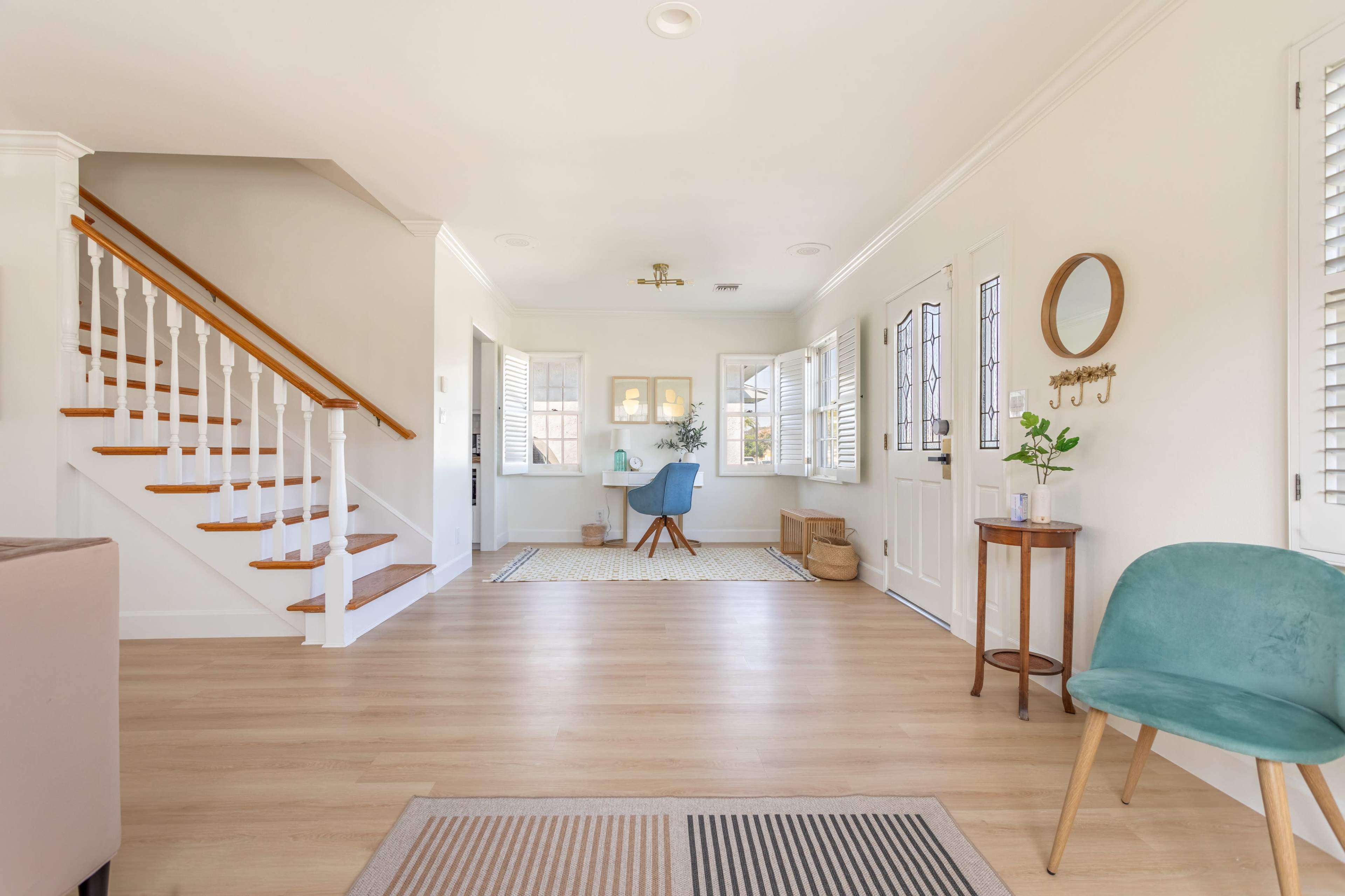 The image shows a bright and airy entryway with a staircase, a small desk in a nook, a circular mirror, and minimal furnishings.
