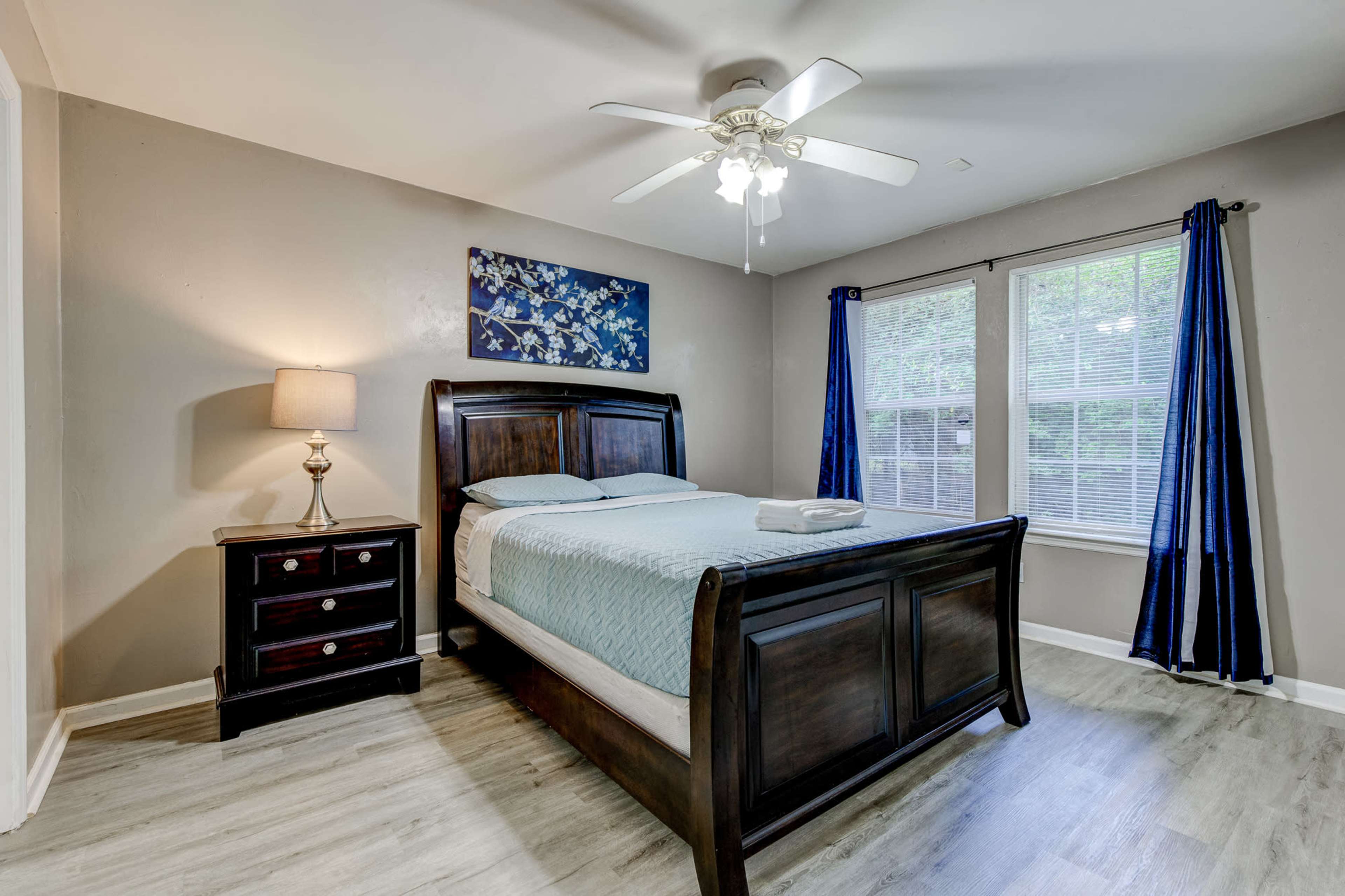 A bedroom featuring a wooden sleigh bed, a nightstand with a lamp, and two windows with blue curtains.