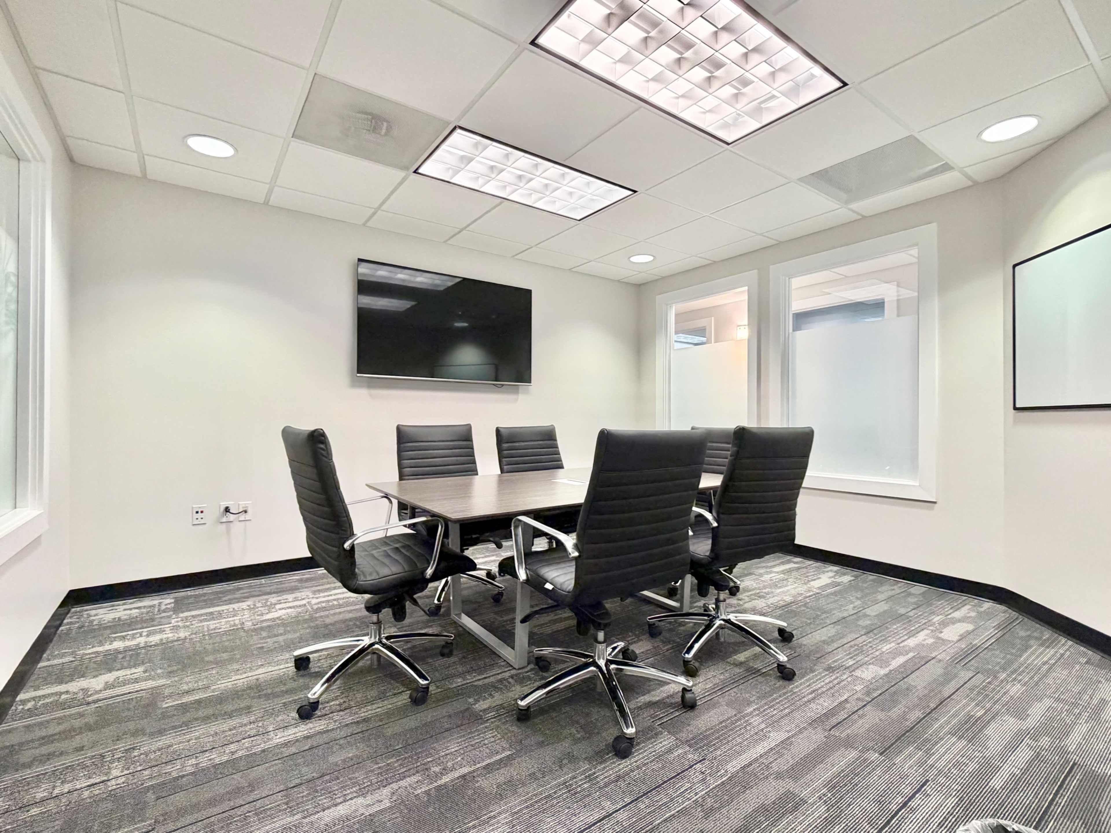 The image shows a modern conference room with a large table surrounded by six black chairs, and a wall-mounted screen.