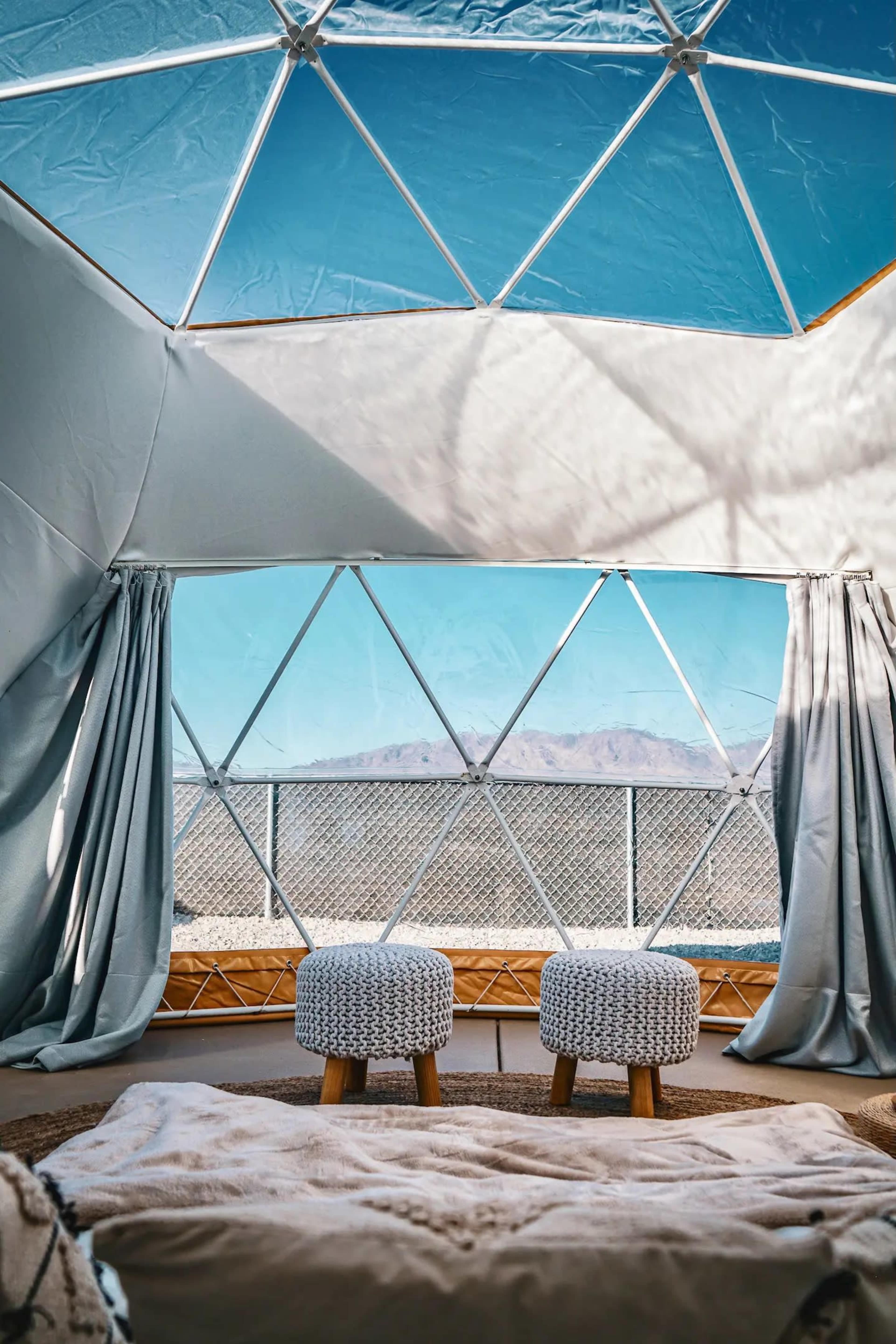 The interior of a geodesic dome features two cushioned stools and a cozy rug, with large windows offering a view of distant mountains under a clear blue sky.
