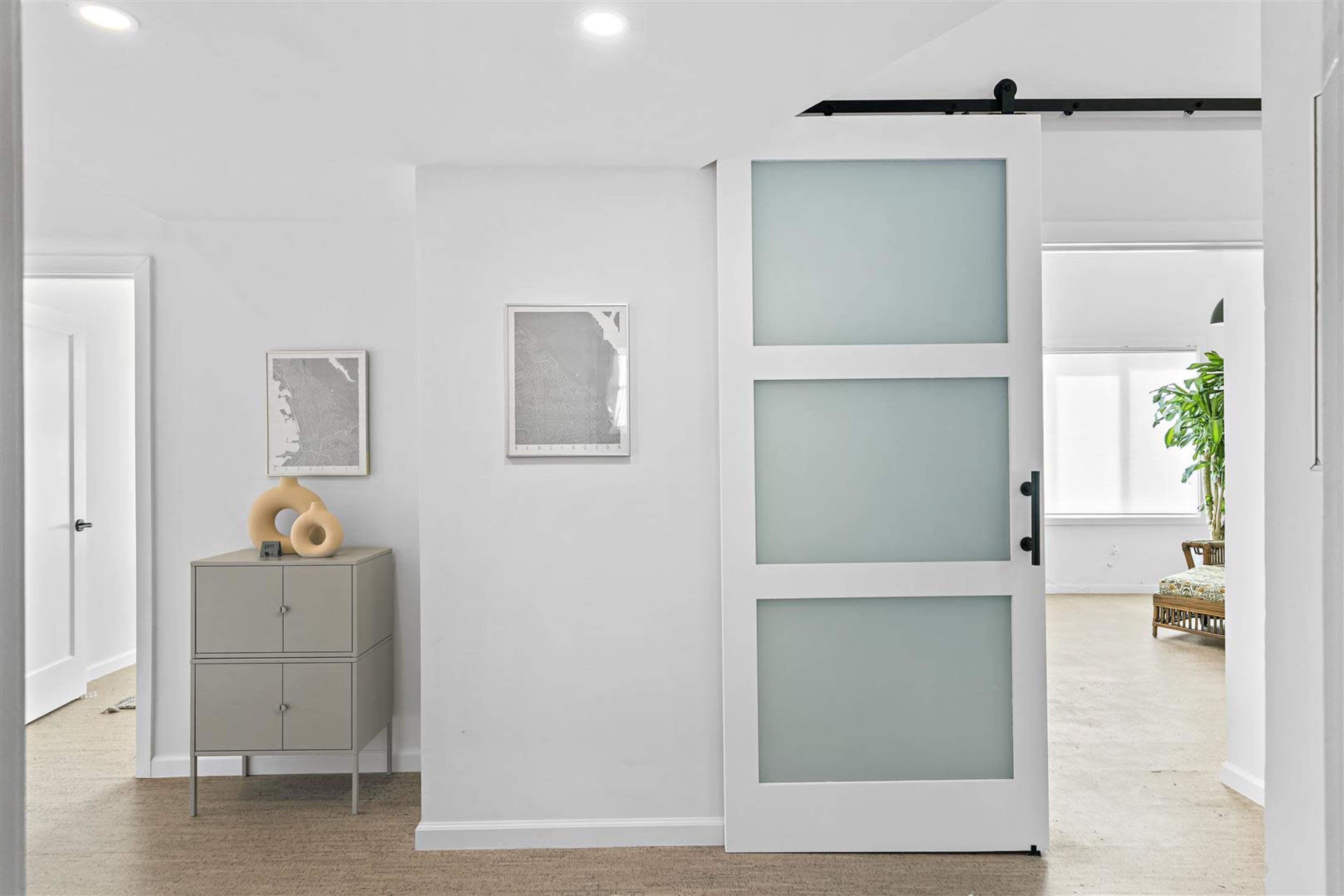 A modern, light-colored hallway with a sliding glass door, a gray cabinet, and framed artwork on the walls.