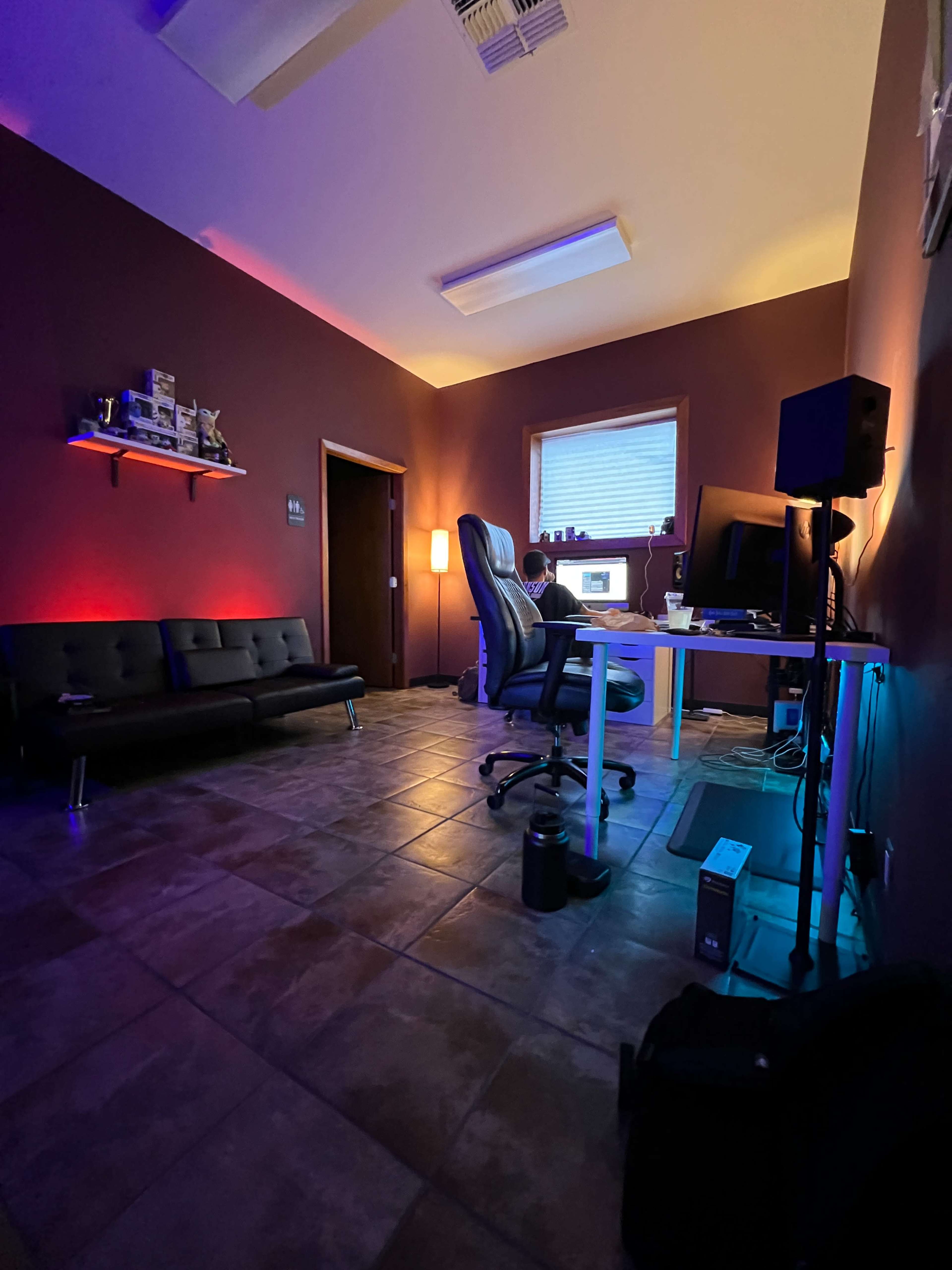 A dimly lit room features a seating area with a black couch, a desk with a computer set up, and ambient light along the walls.