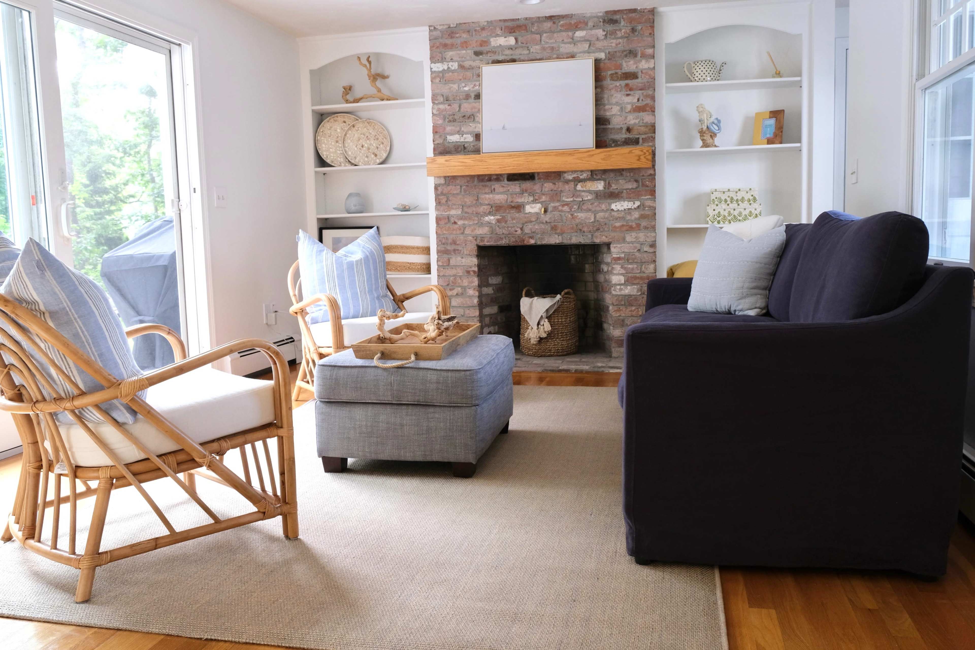 A cozy living room with a brick fireplace, a blue upholstered chair, a light gray ottoman, and two rattan chairs arranged around a neutral-colored area rug.