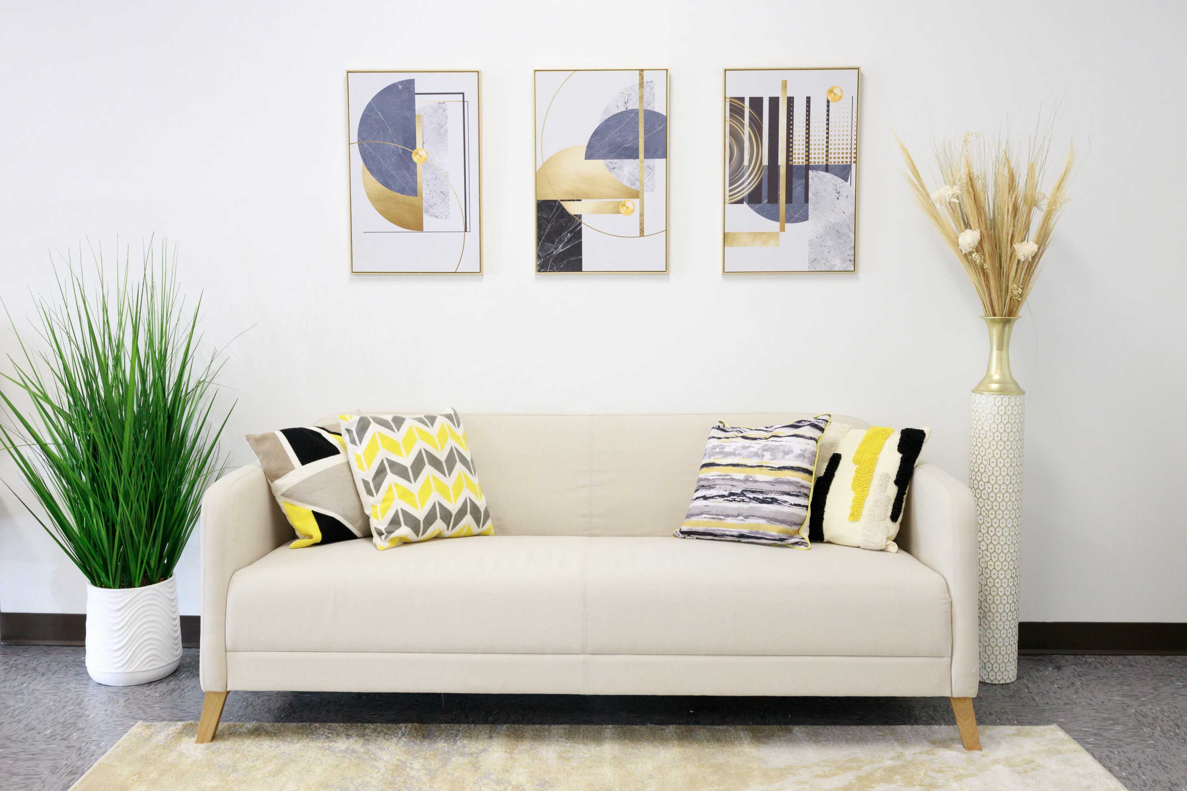 A modern living room features a light-colored sofa with patterned pillows, flanked by tall potted plants and decorative wall art.