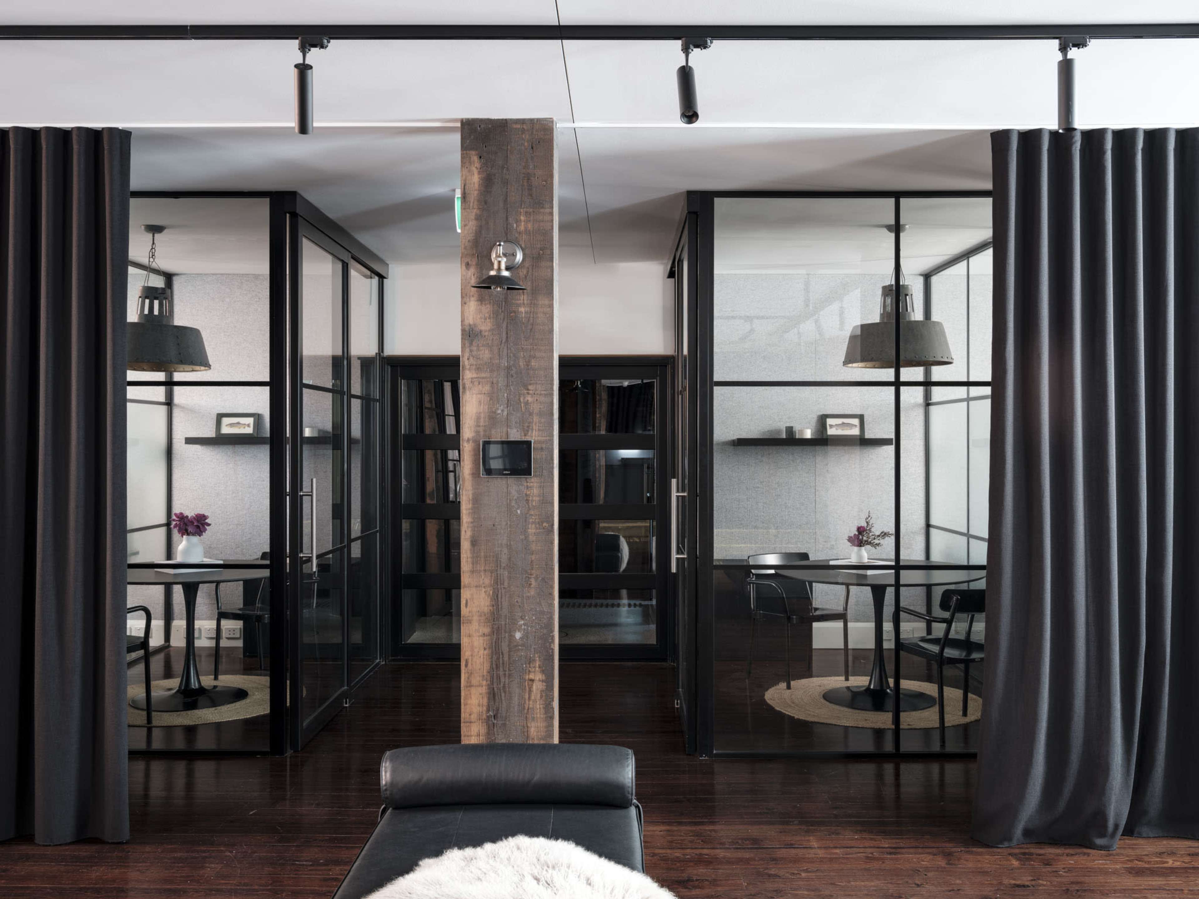 The image shows a modern office space featuring two glass-enclosed meeting rooms separated by a wooden post and dark curtains, with minimalist furniture and decoration.