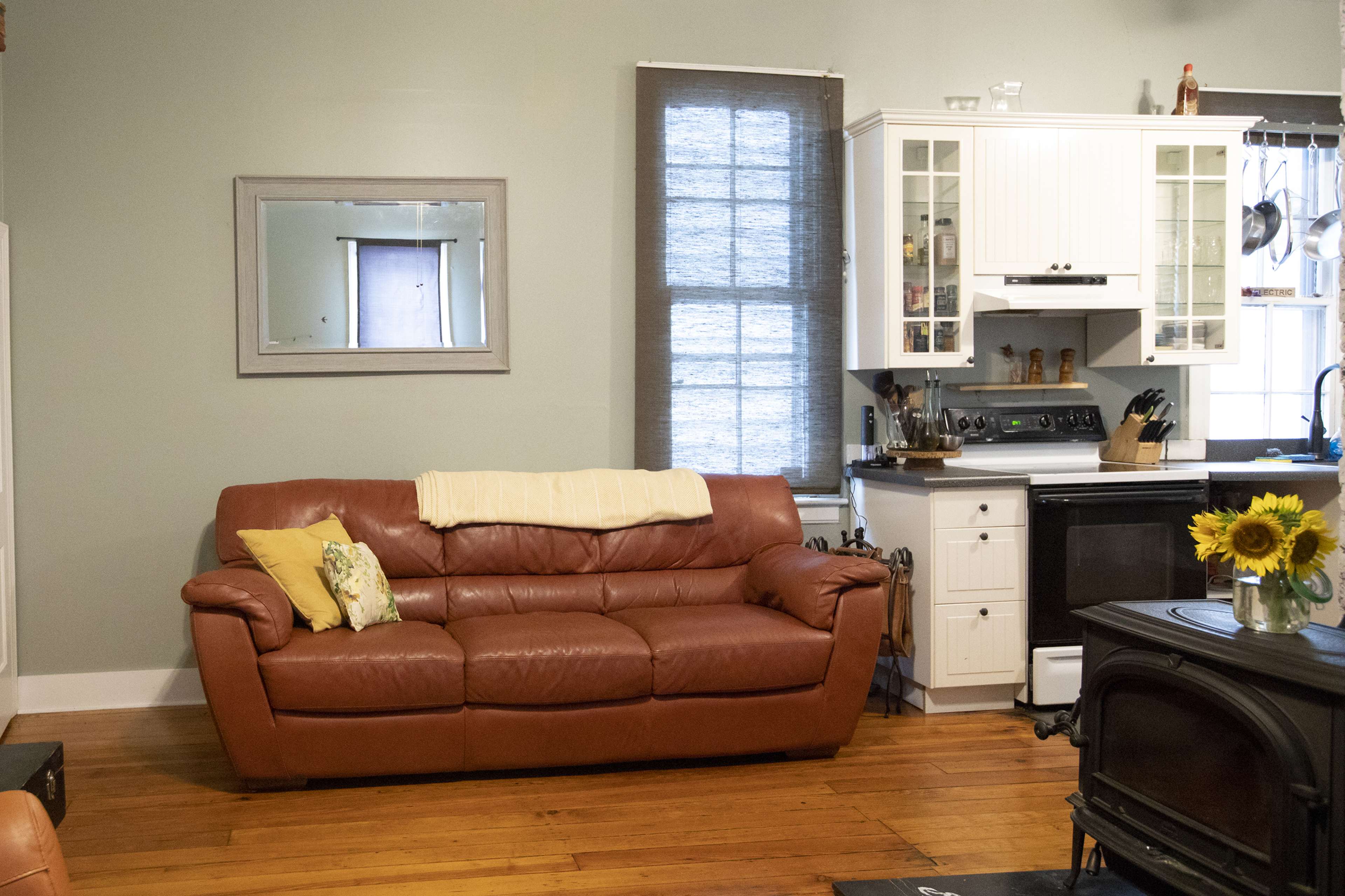 A living room with a brown leather couch, a small coffee table with sunflowers, and a modern kitchen area visible in the background.