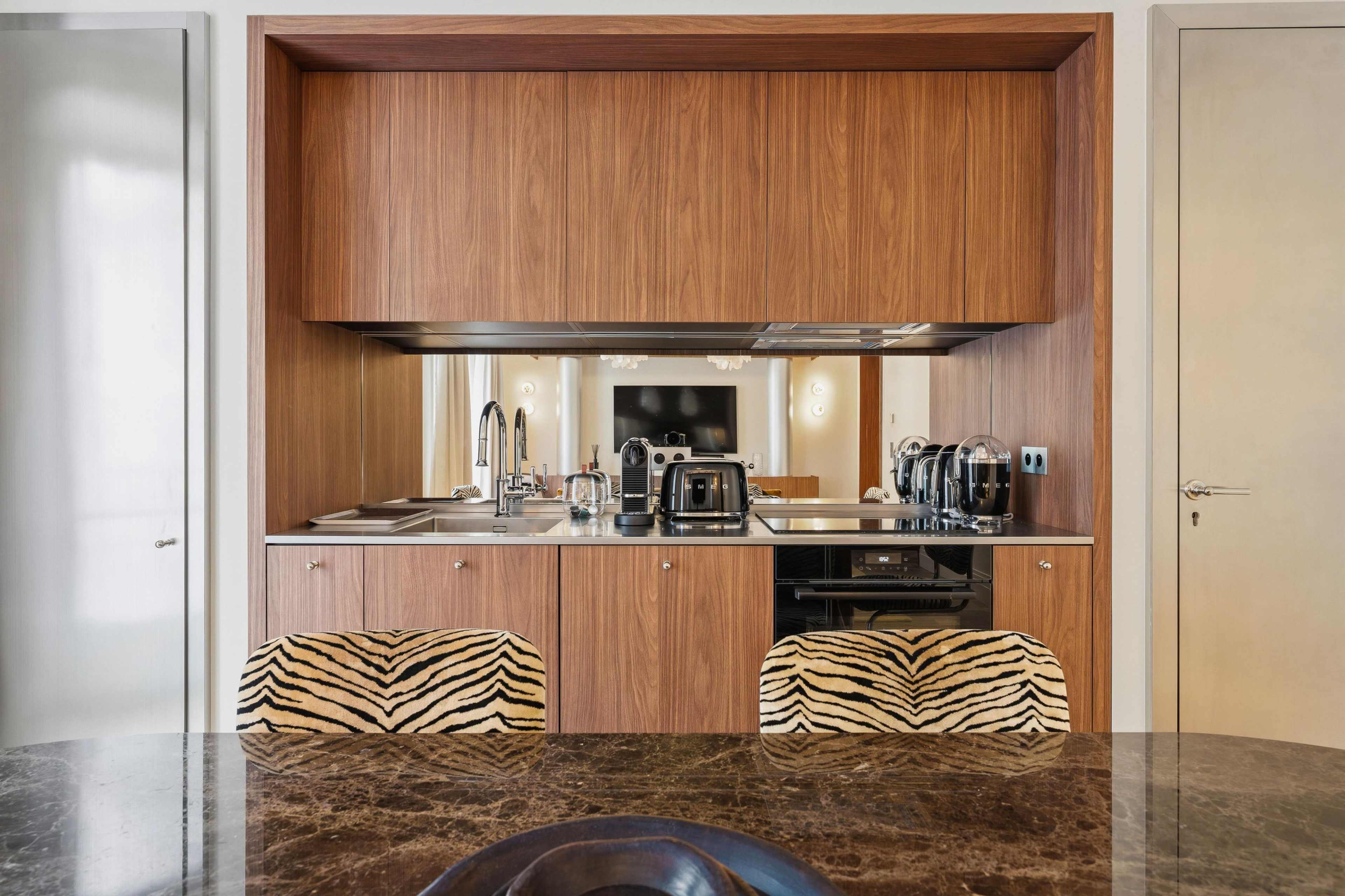 The image shows a modern kitchen area with wooden cabinetry, stainless steel appliances, and two zebra-striped chairs at a marble dining table.