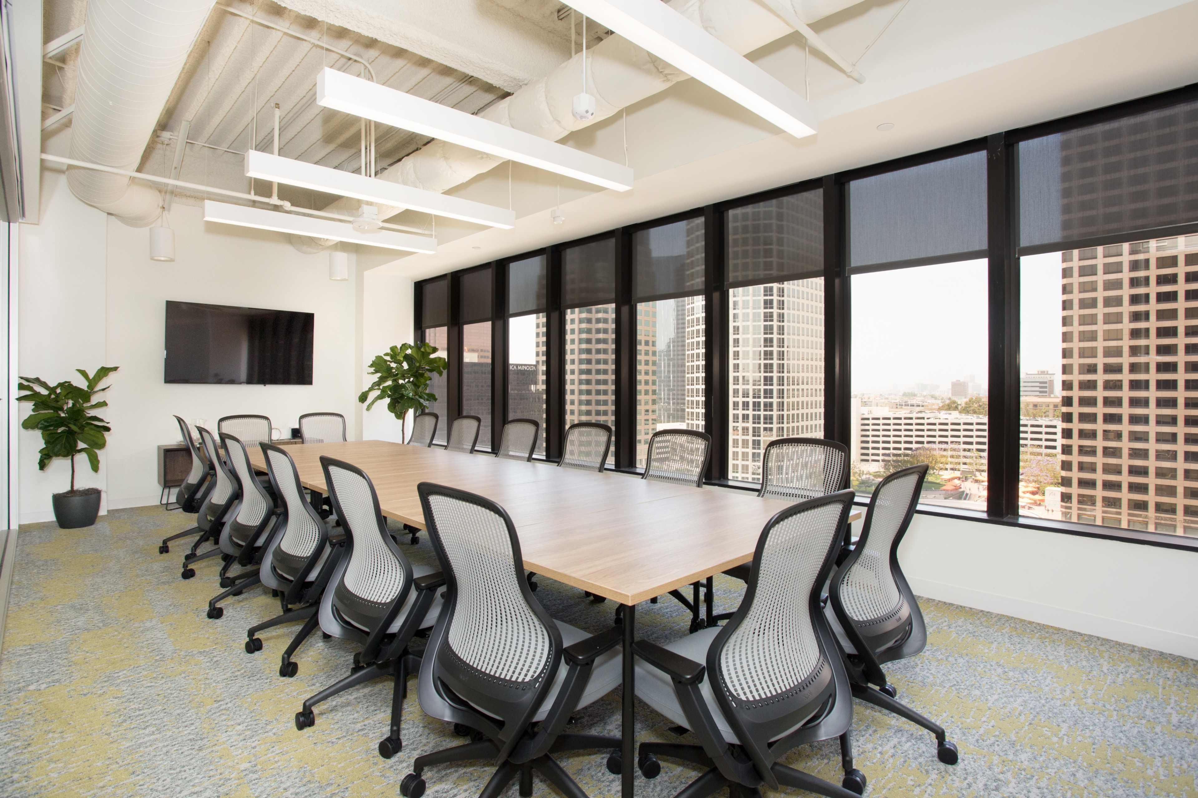 A conference room features a long wooden table surrounded by ergonomic chairs, with large windows offering a view of a city skyline.
