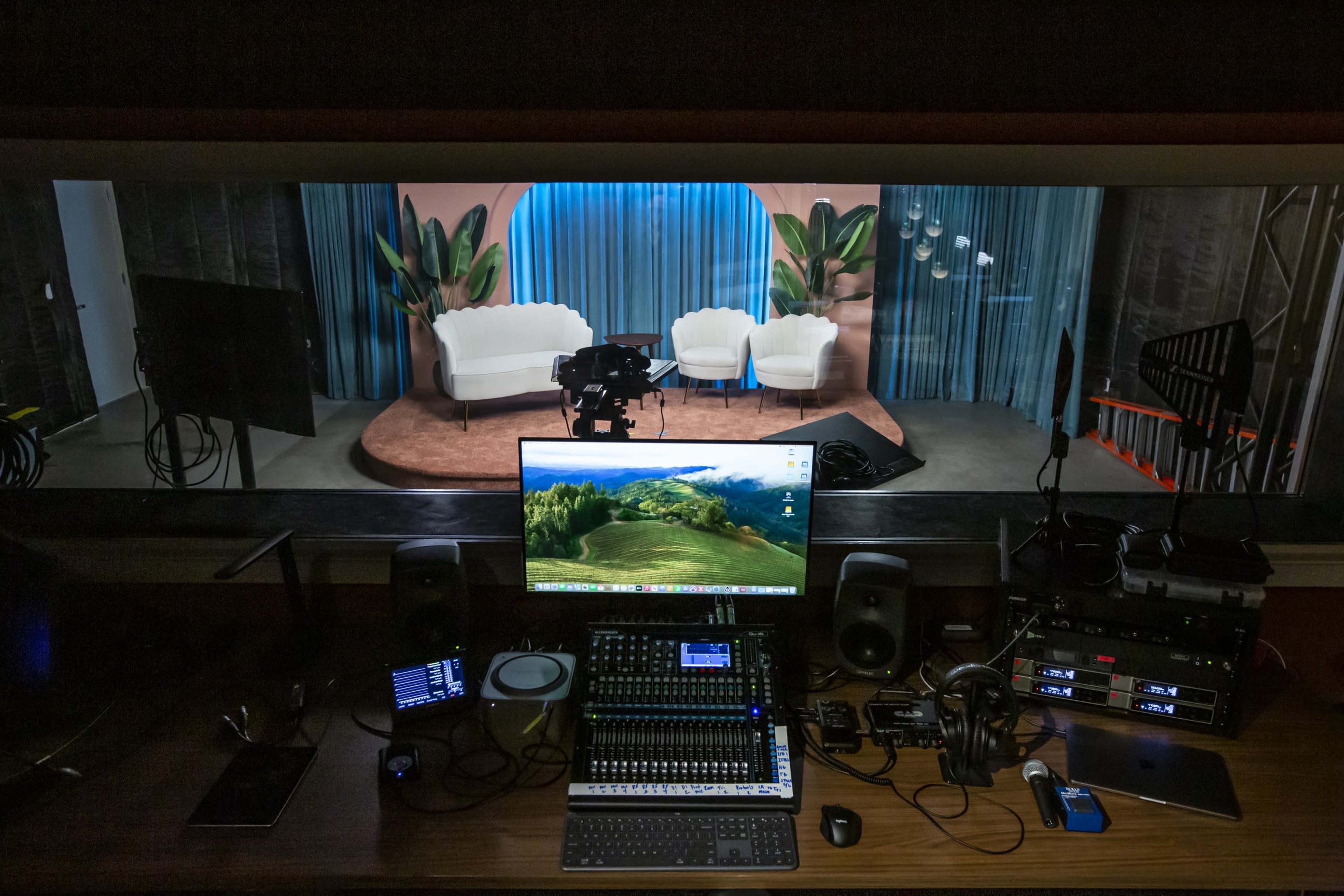 A broadcasting control room features a large monitor, audio equipment, and a view of a brightly lit studio set with two white chairs and a backdrop of curtains and plants.