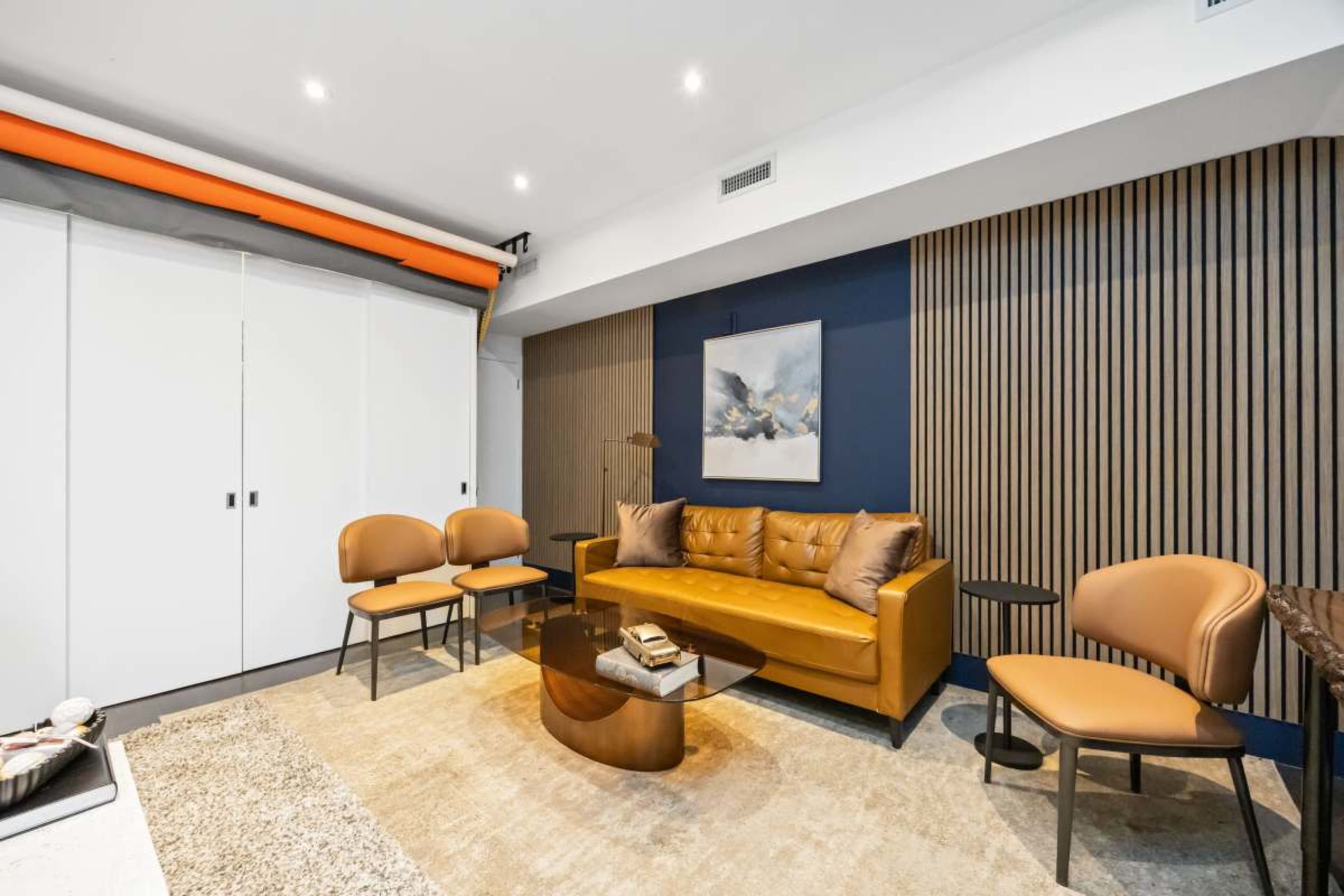 The image shows a modern living room featuring a yellow-orange sofa, two matching chairs, a coffee table, and a large piece of wall art against a dark blue accent wall.