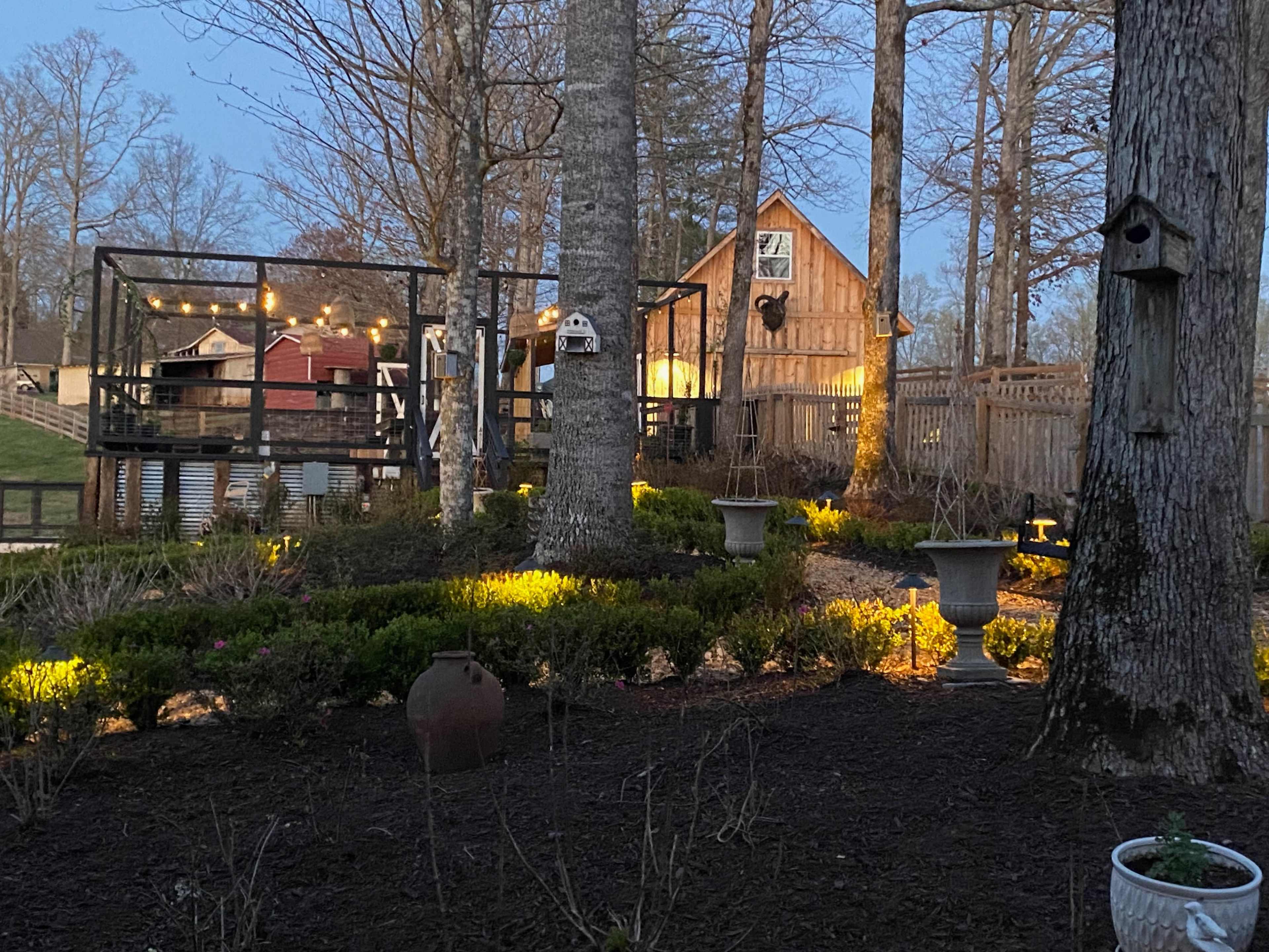 A landscaped garden area featuring stone paths, birdhouses, and a wooden cabin in the background, illuminated by string lights among the trees.