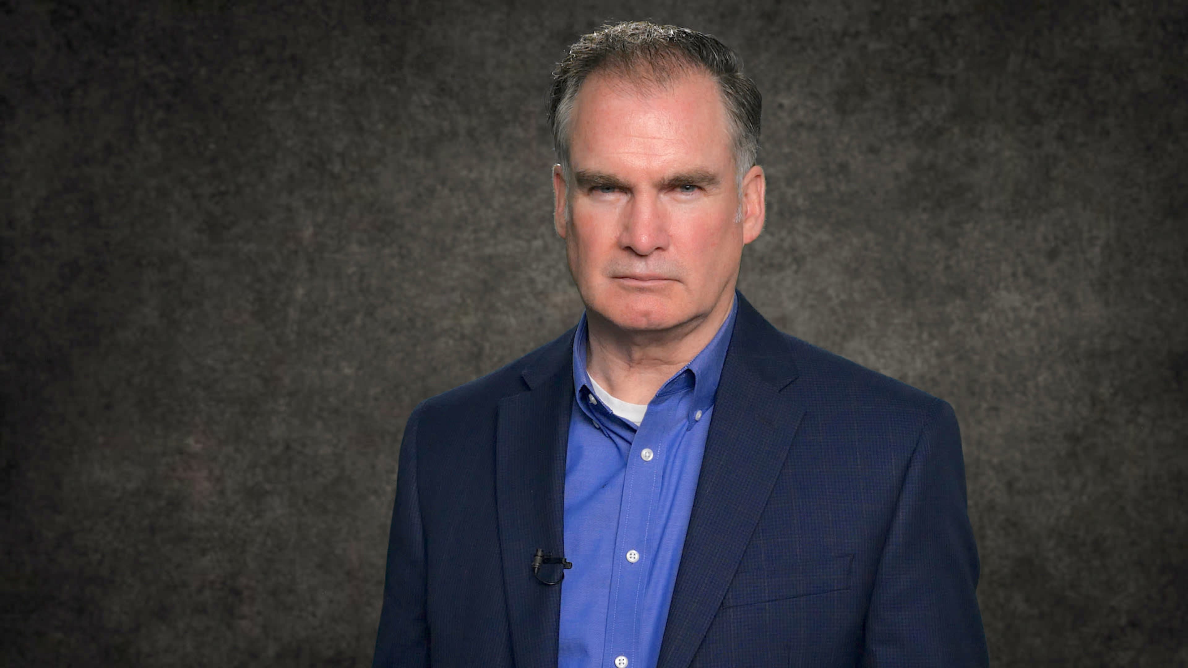 A man in a dark blazer and light blue shirt stands in front of a textured gray backdrop.