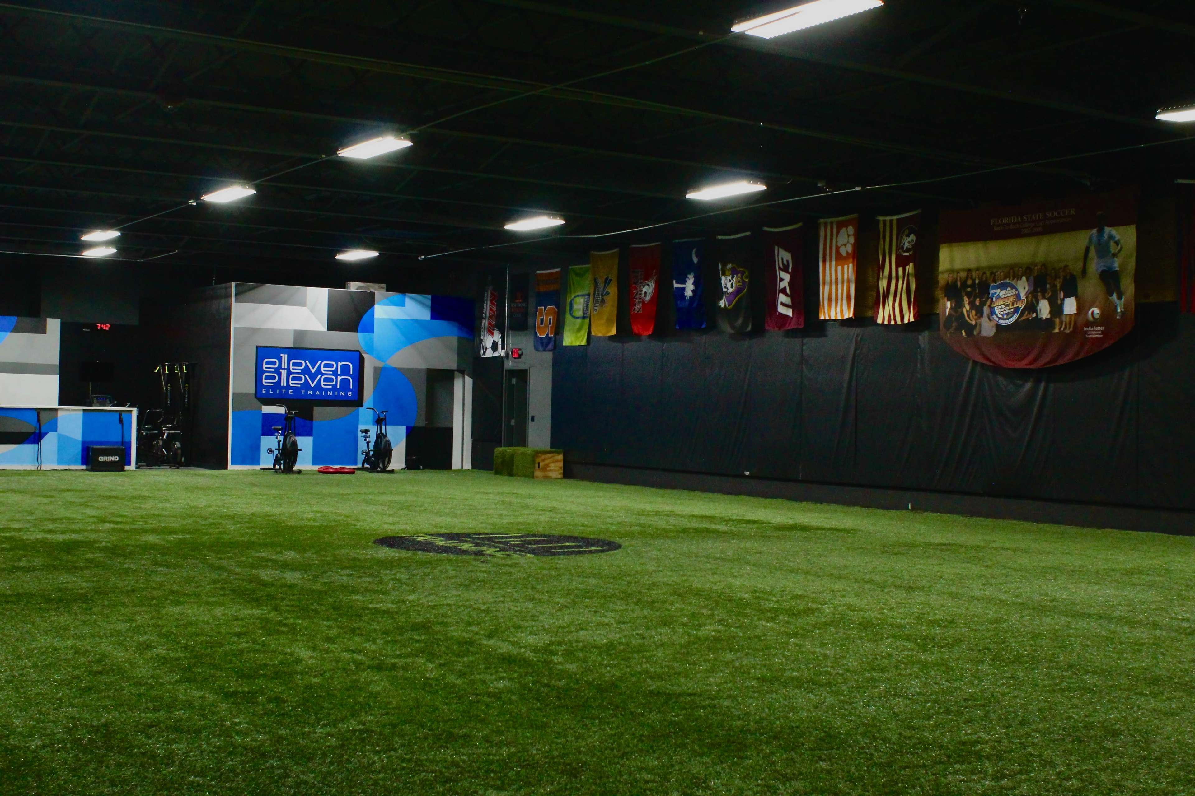 An indoor training facility featuring a turf field, a service area with equipment, and various flags hanging on the walls.