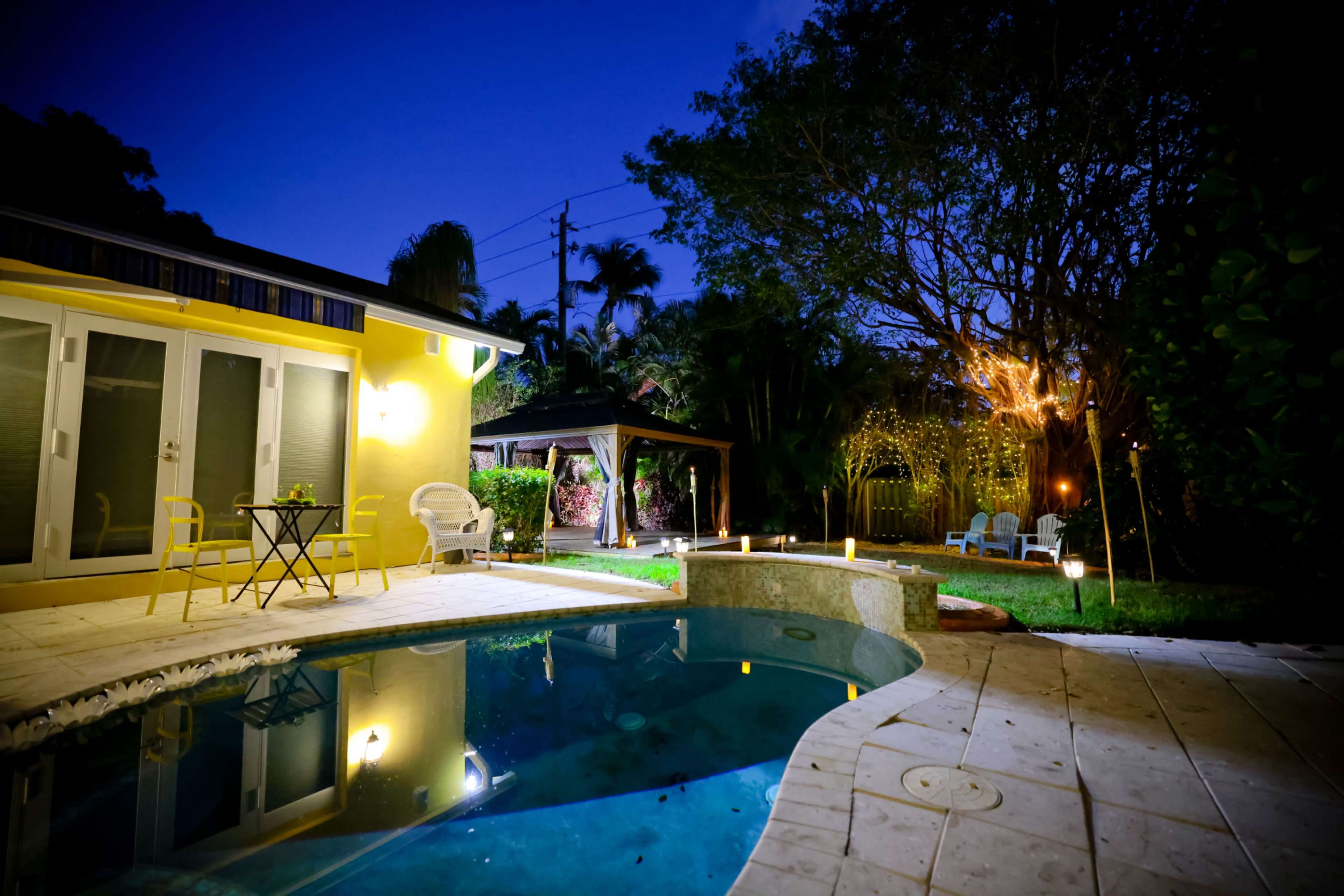 The image shows a lit outdoor area featuring a pool, a small table with chairs, and a gazebo surrounded by greenery at dusk.