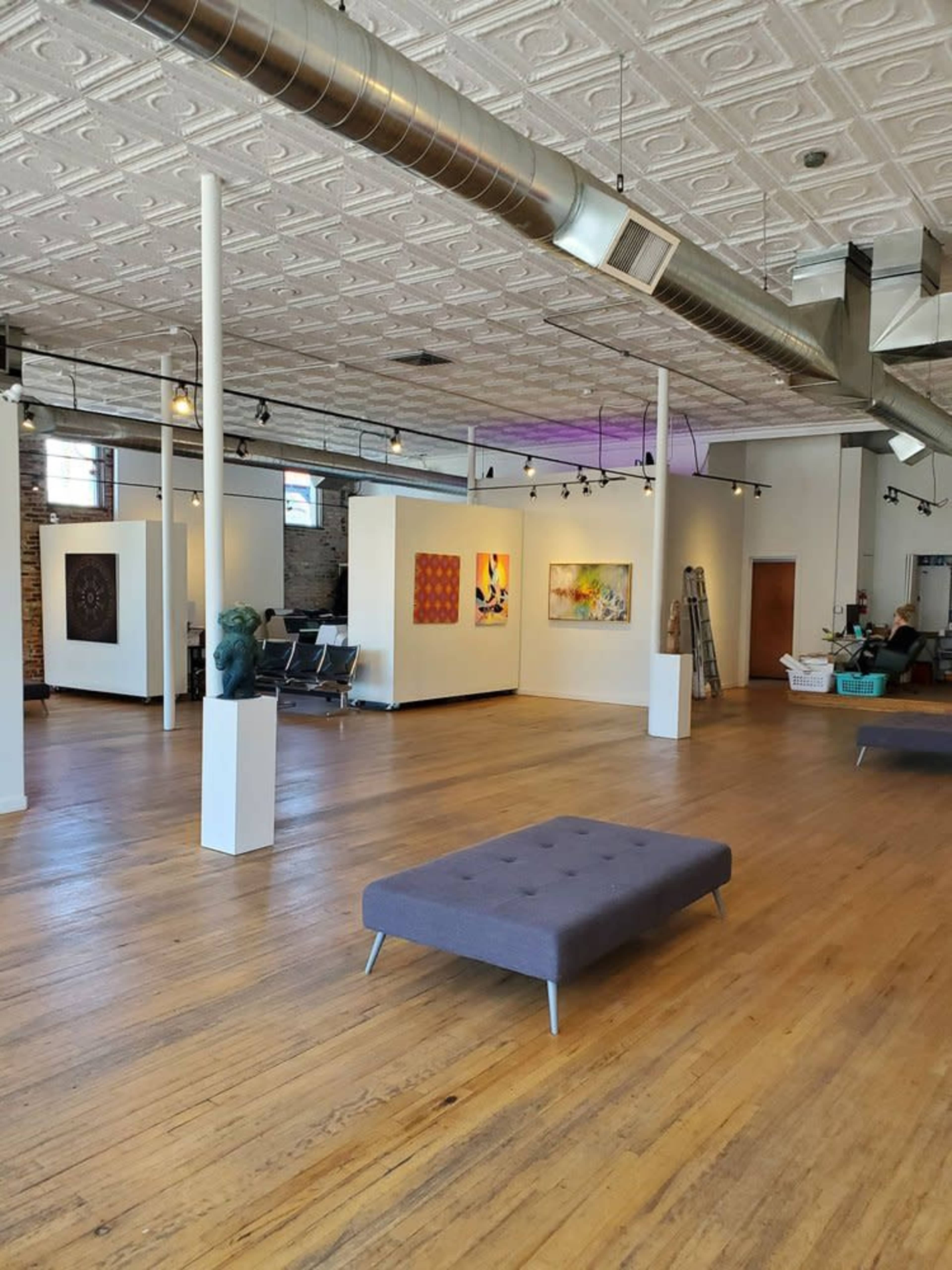 The spacious art gallery features white walls adorned with various pieces of artwork, wooden flooring, and a seating area with a gray tufted bench.