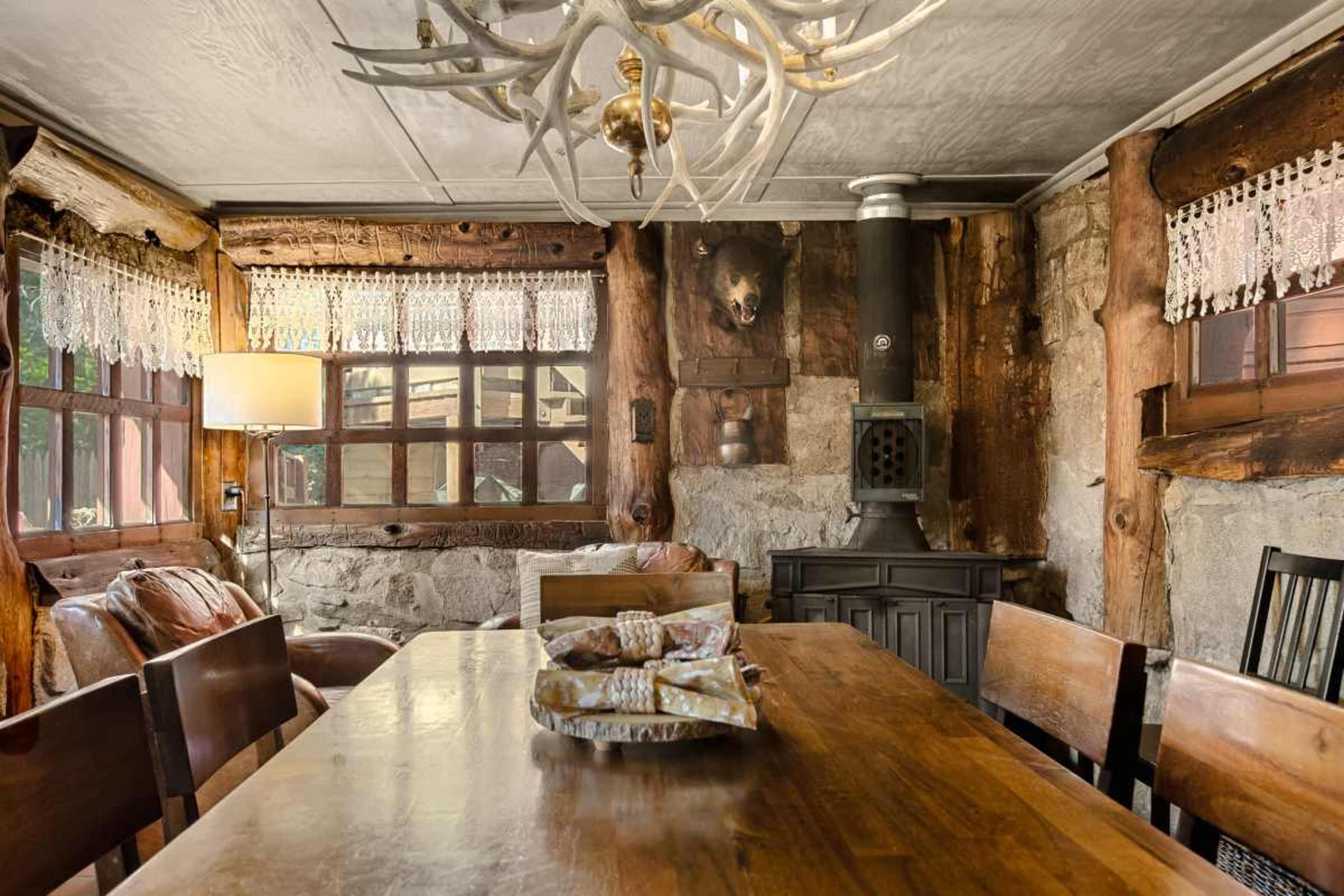 A wooden dining table sits in a rustic room with stone walls, adorned with wooden beams and an antler chandelier overhead.