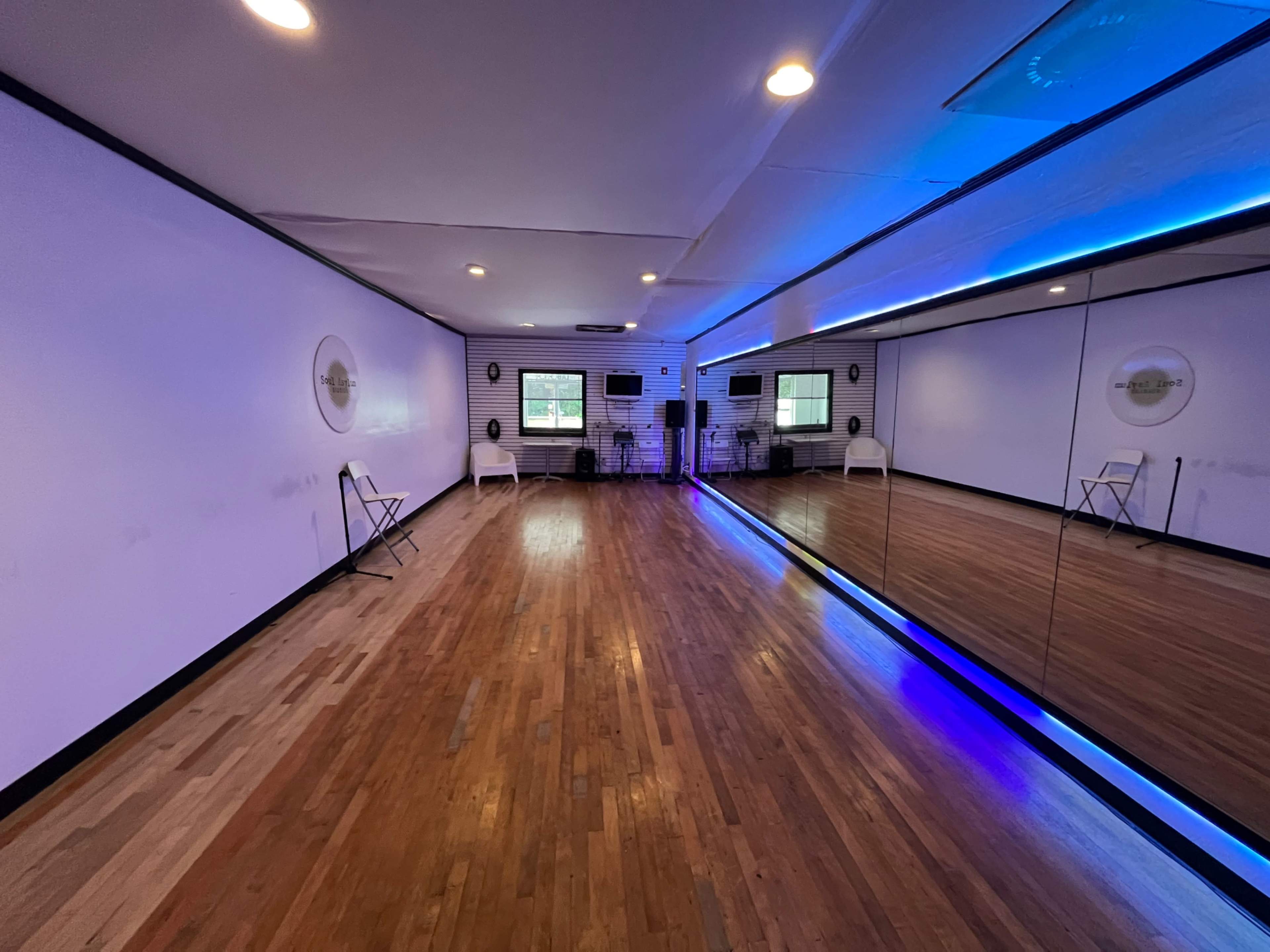 A spacious dance studio features hardwood floors, white walls, and a large mirror with blue lighting along one side.