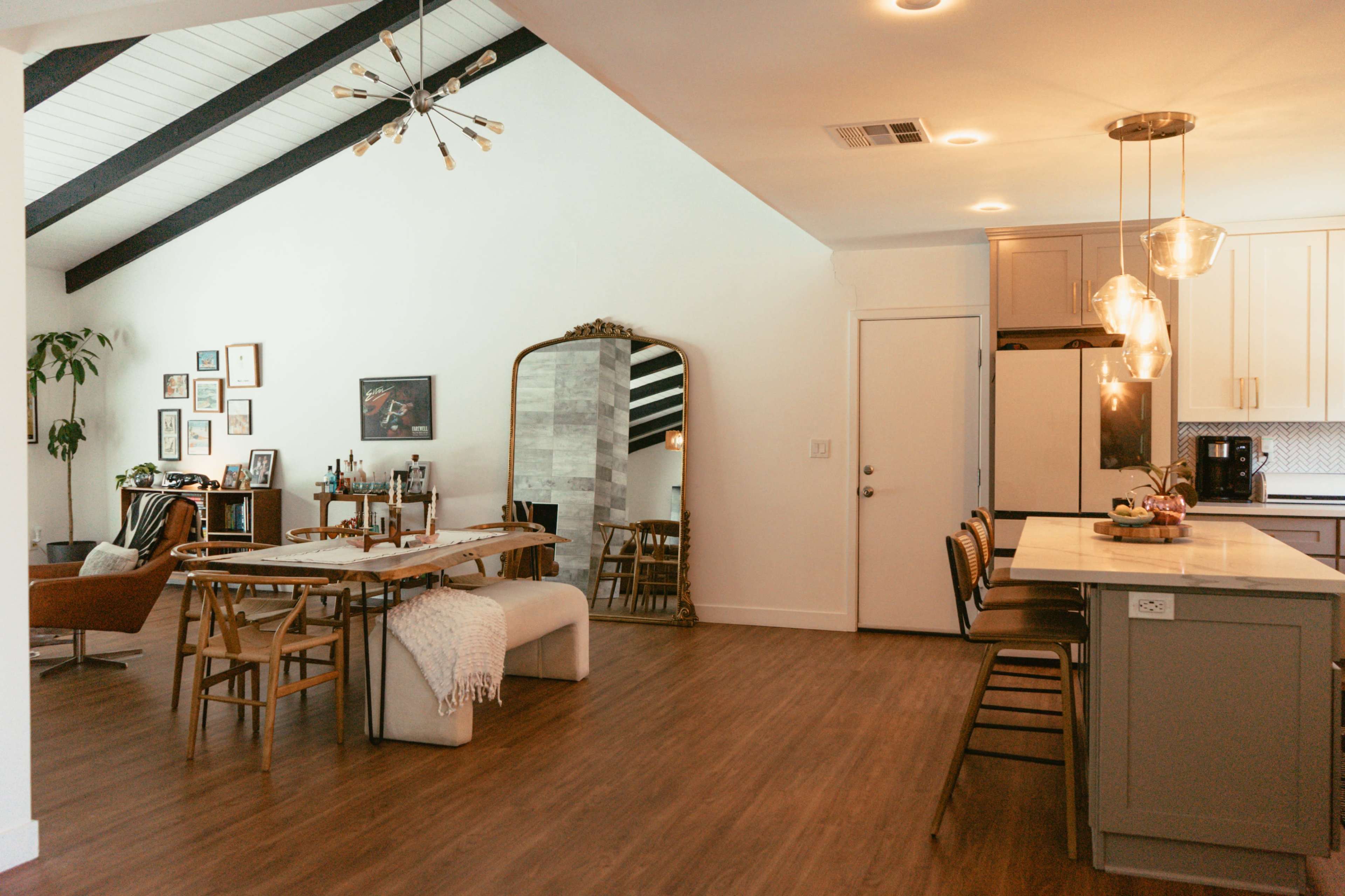 The image shows a modern open-concept living space featuring a dining area with wooden chairs, a kitchen with an island, and a cozy seating area with a large mirror and art on the walls.
