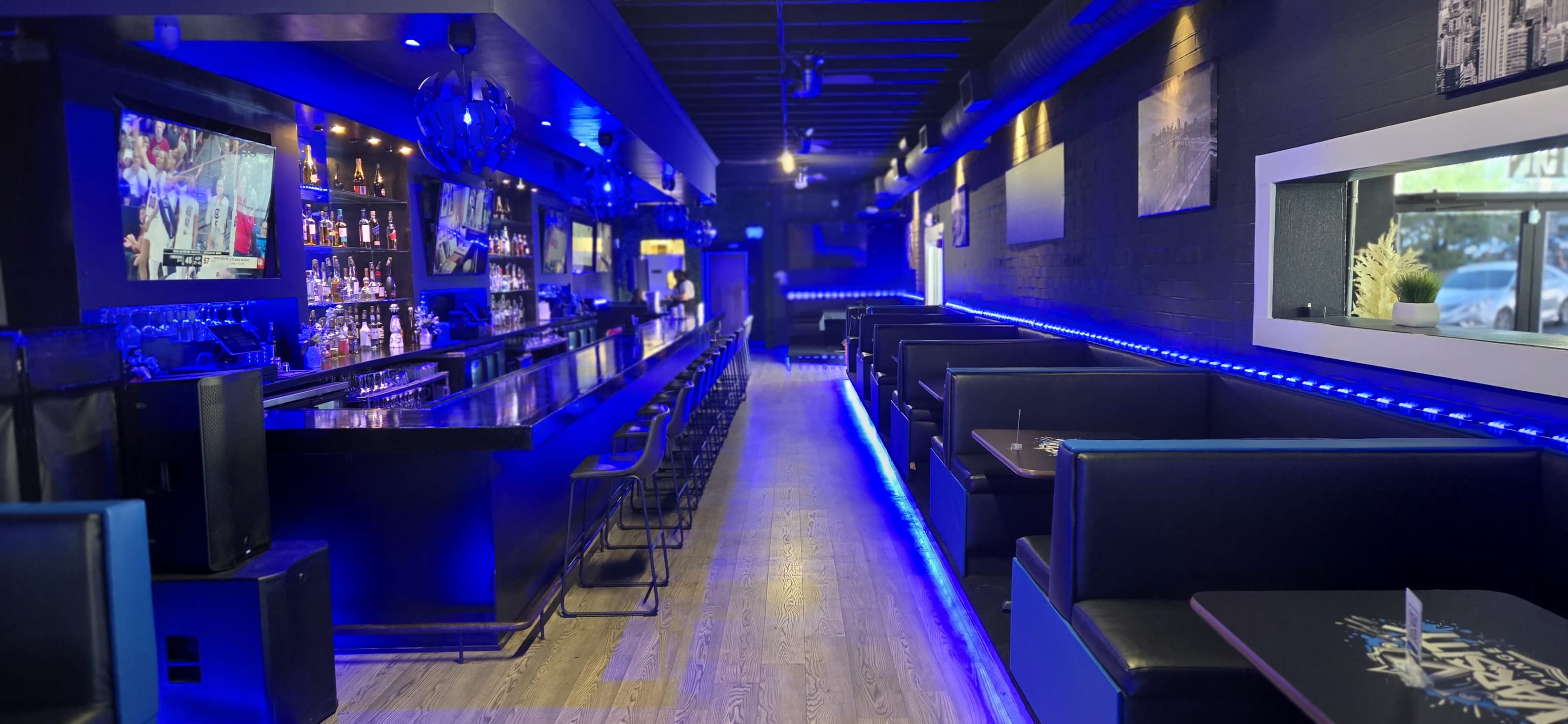 The image shows a modern bar interior with blue LED lighting, wooden flooring, and a long counter with bar stools on one side, flanked by booths along the walls.
