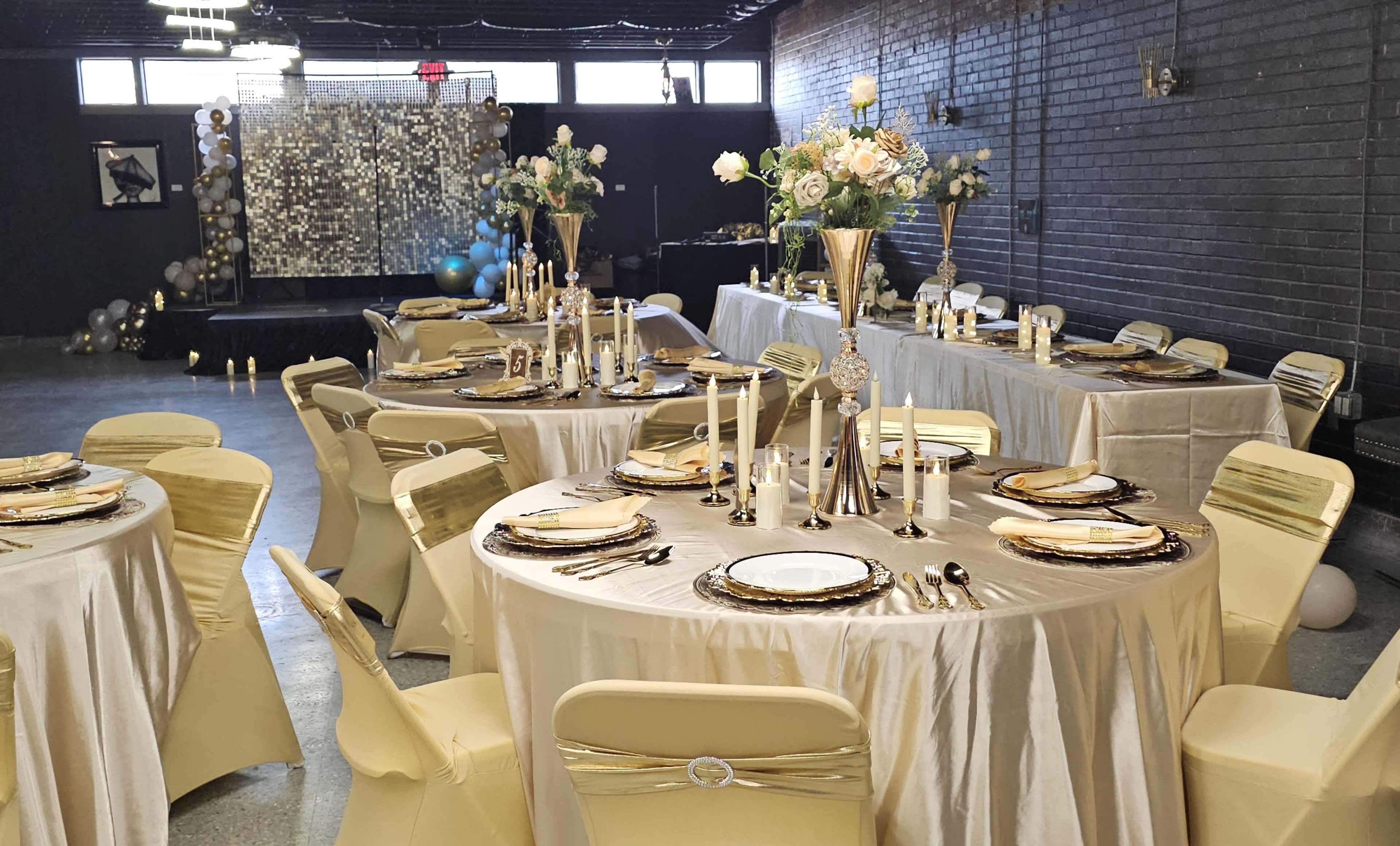 The image shows a beautifully arranged banquet hall with round tables covered in elegant gold and cream tablecloths, each set with fine dining plates, silverware, and tall candle holders, while a decorative backdrop adorned with flowers and balloons is visible in the background.