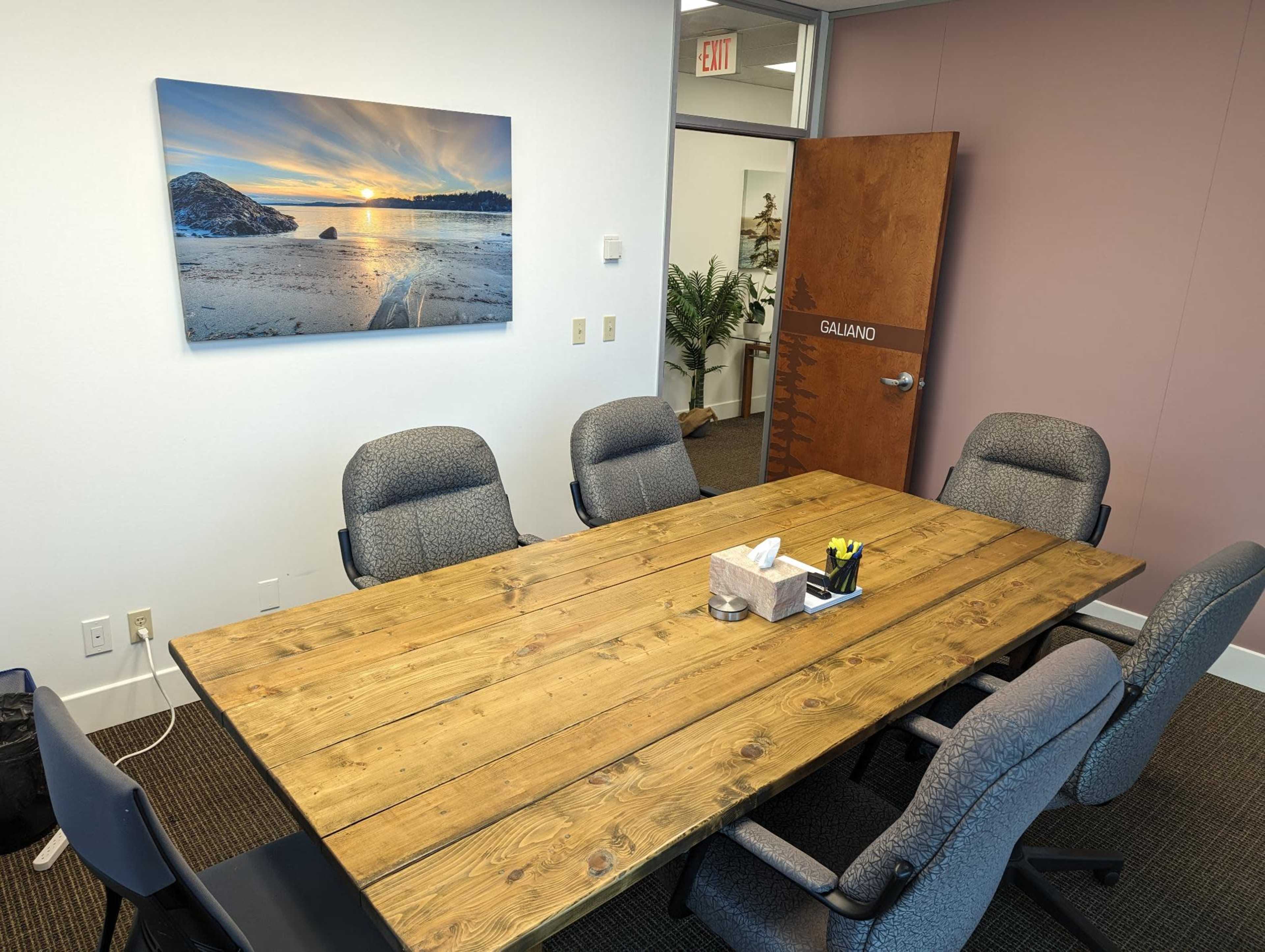 A wooden conference table surrounded by gray office chairs is set in a meeting room with a landscape photograph on the wall.