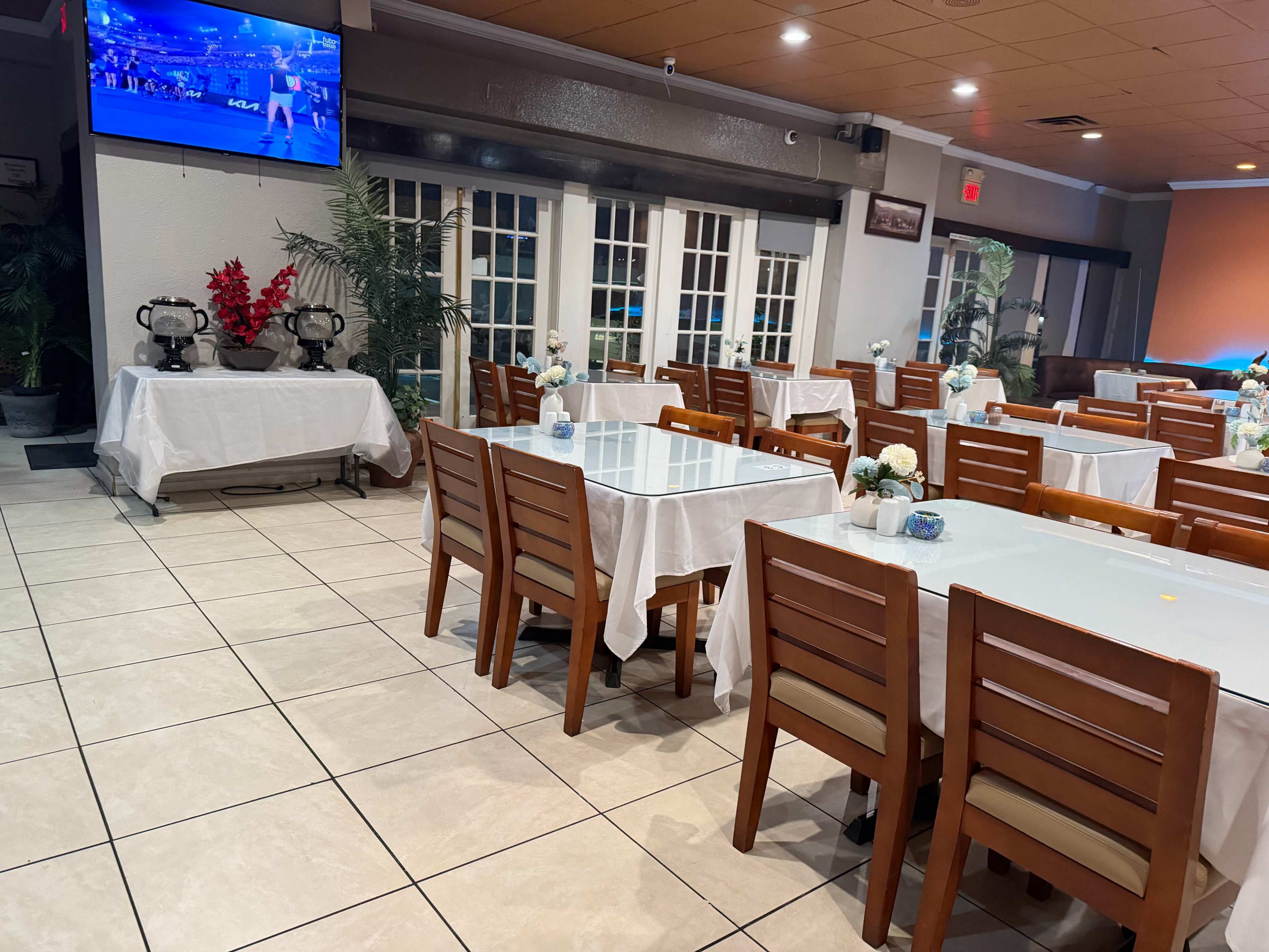 The image shows an empty restaurant interior with several tables covered in white tablecloths, wooden chairs, and a television displaying a sports event on the wall.