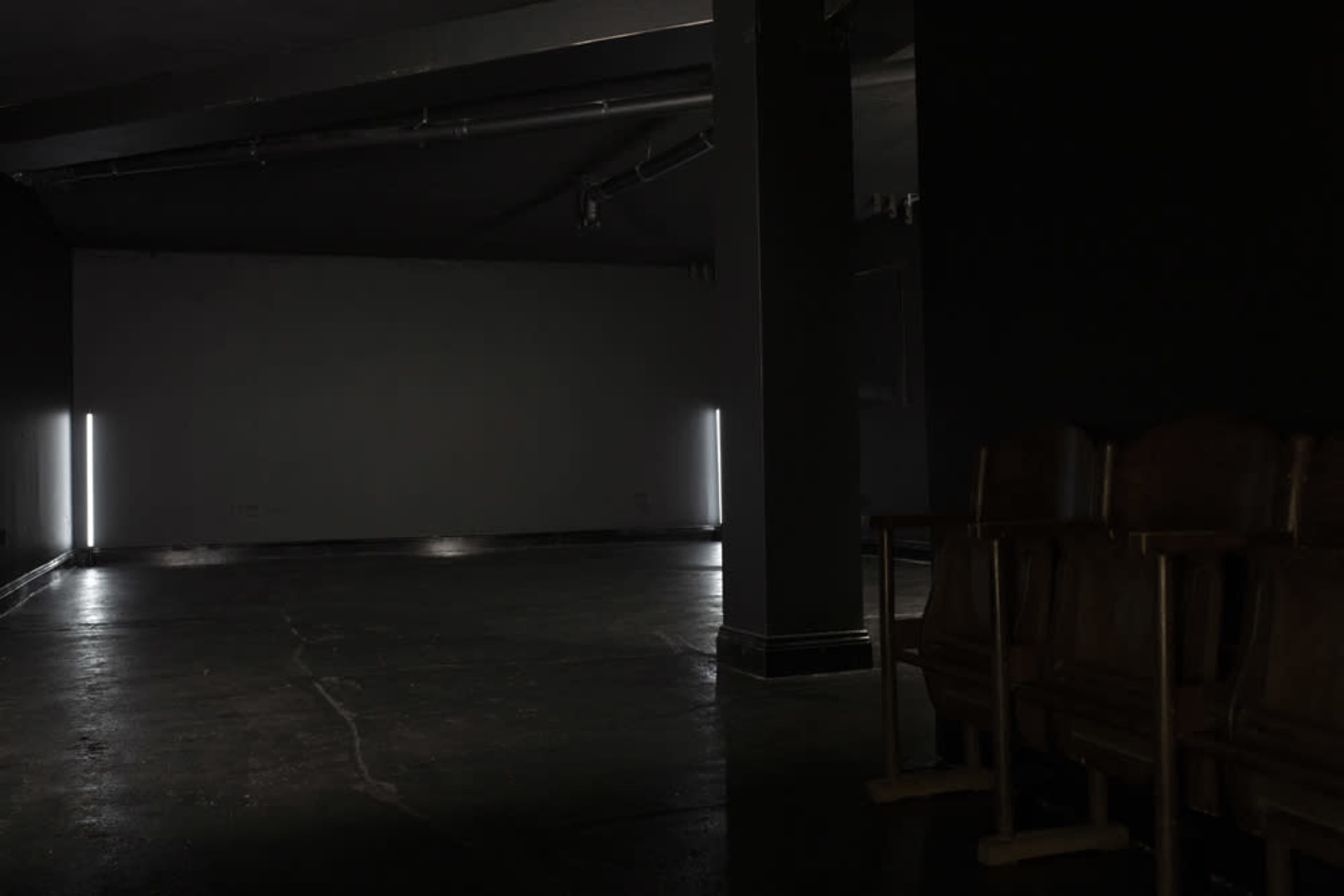 A dimly lit room features two vertical light fixtures on the walls and a row of wooden chairs along one side.