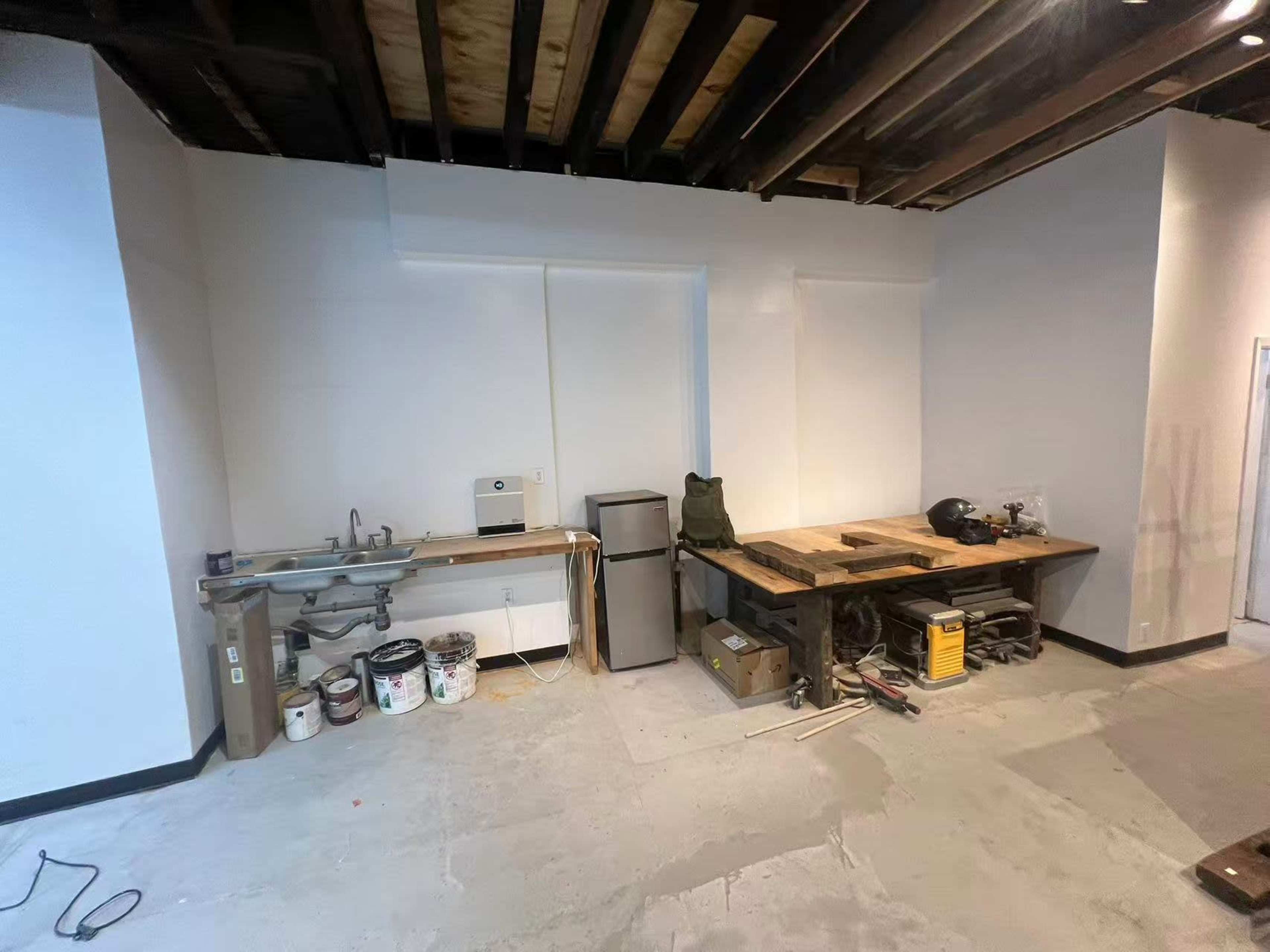 The image shows an unfinished room with exposed beams on the ceiling, a workbench against one wall, a small refrigerator, and various tools and boxes on the floor.