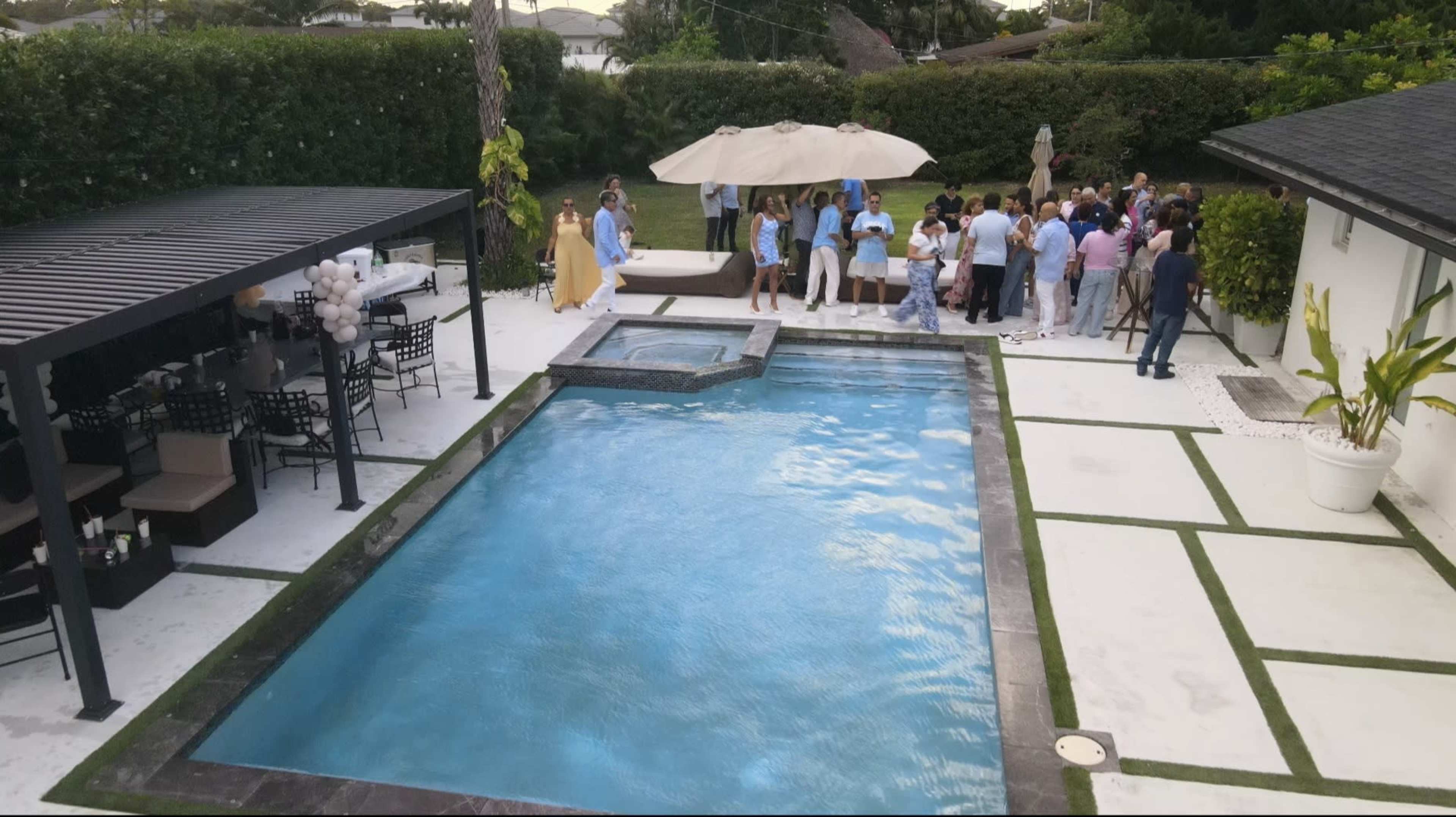 A large group of people gathers around a swimming pool in a residential backyard, with shaded seating areas and landscaping in the background.