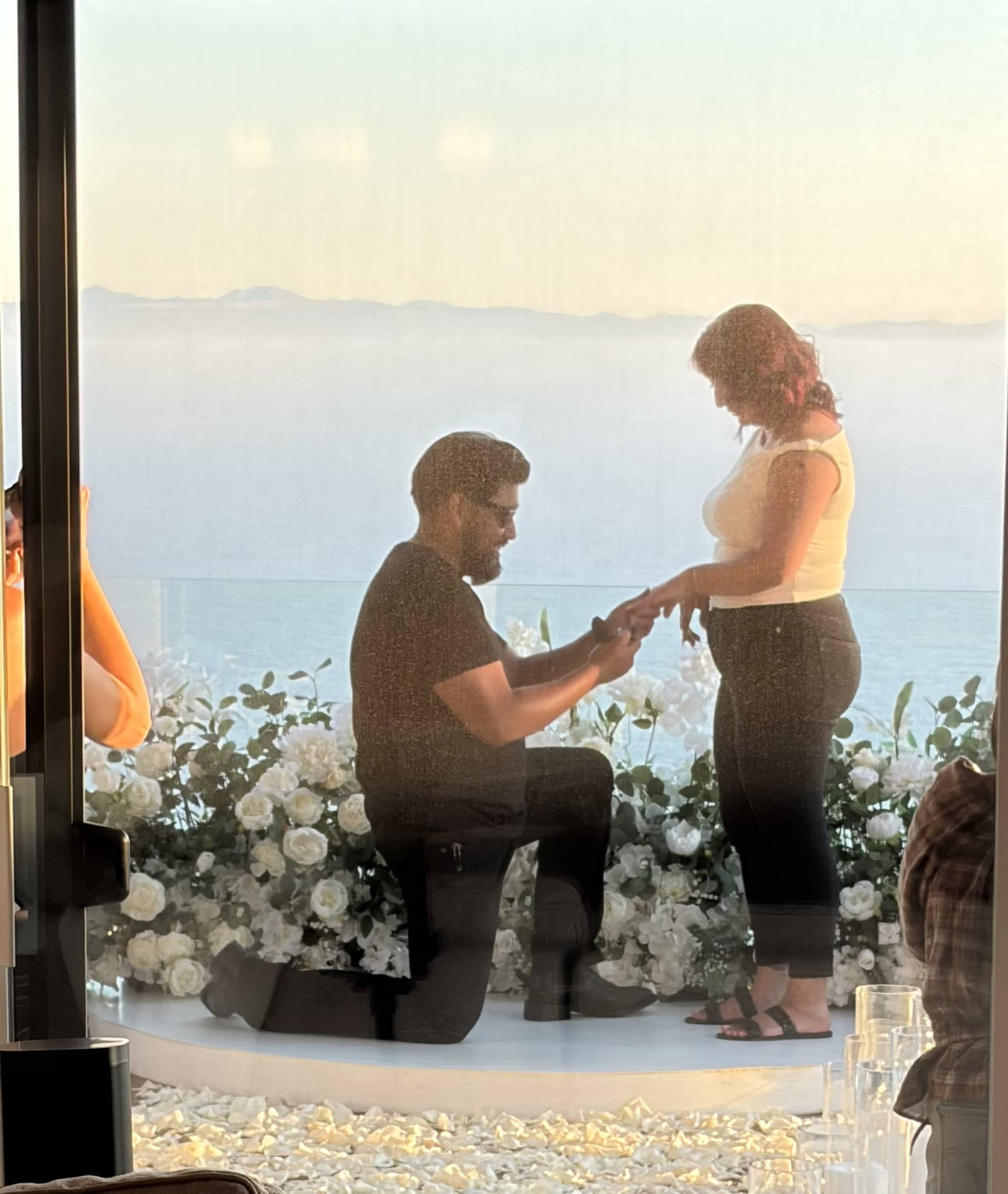 A man proposes to a woman while kneeling in front of a floral display overlooking a scenic view.