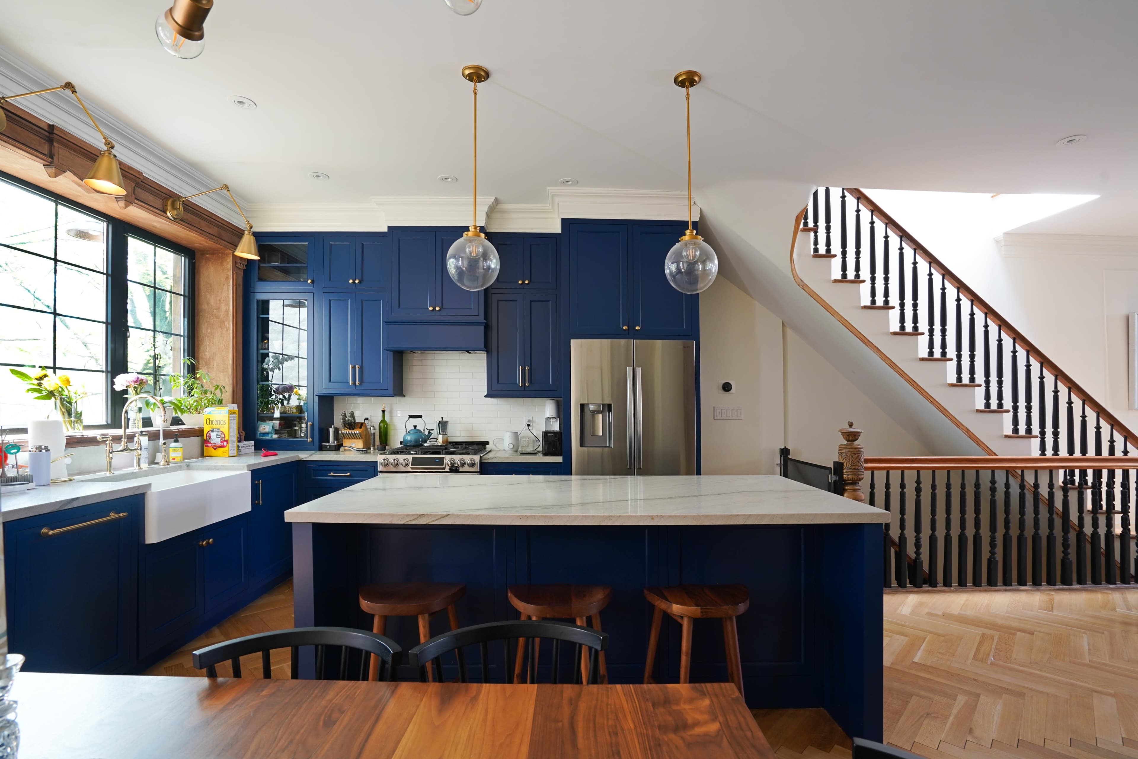 The image shows a modern kitchen with blue cabinetry, a large marble island, pendant lighting, and a staircase in the background.