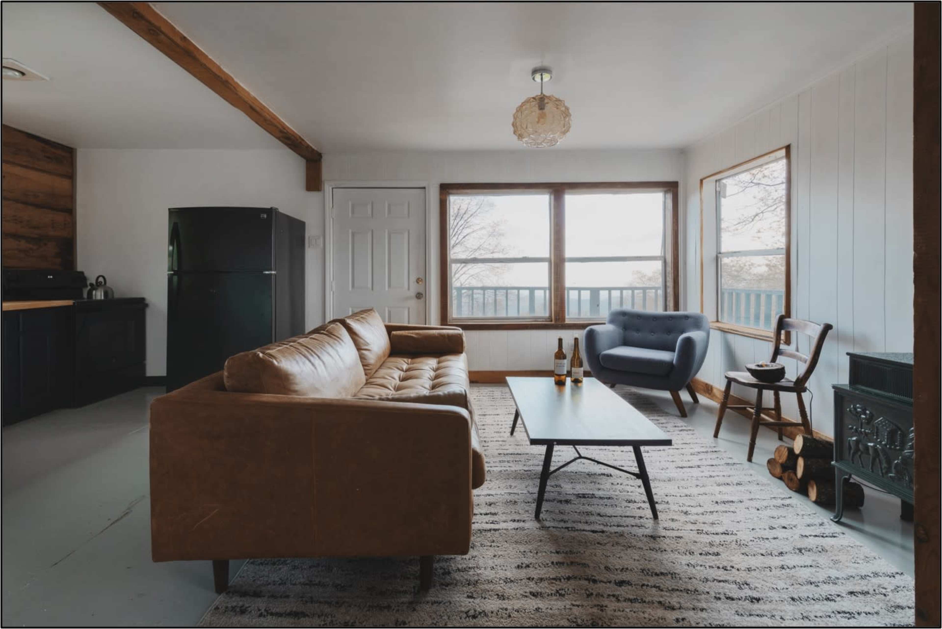 A living room with a brown sofa, a blue armchair, a coffee table, and a view out of large windows, featuring a black refrigerator and logs stacked next to a fireplace.