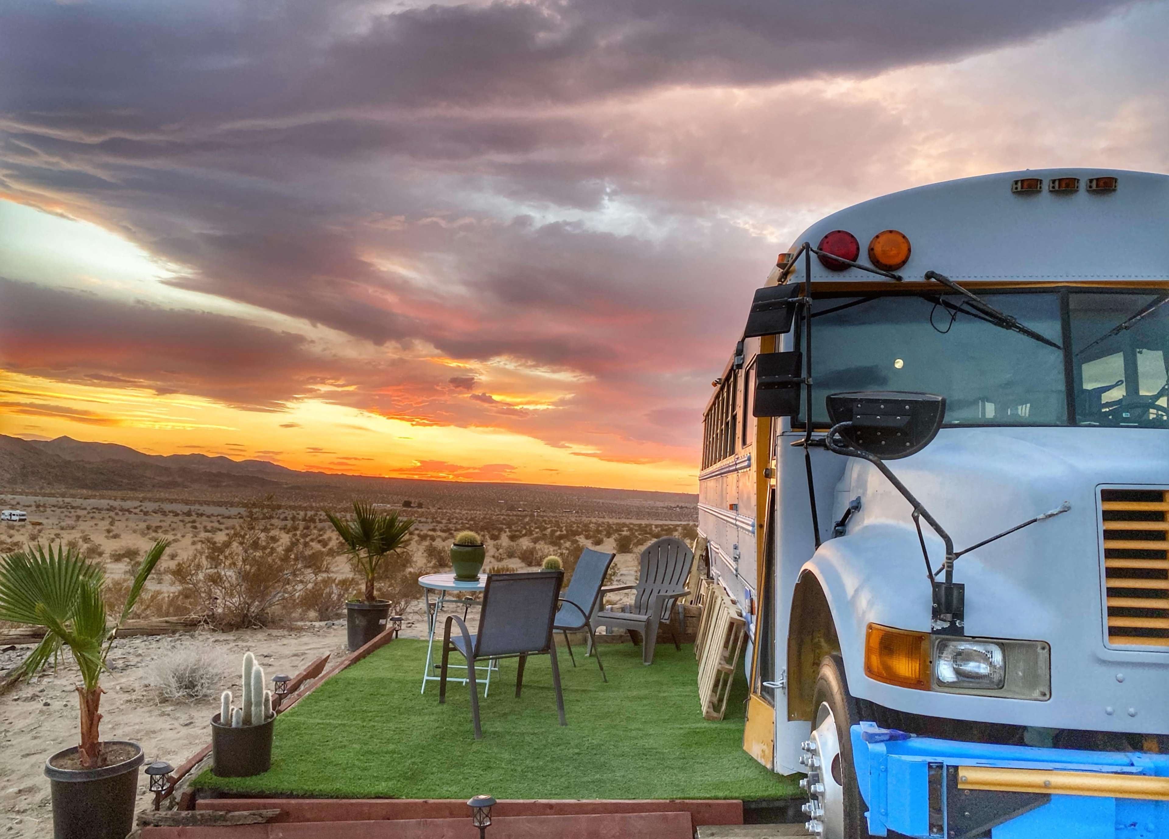 A vintage bus is parked on a raised platform surrounded by desert scenery, with a table and chairs set up nearby, under a colorful sunset sky.