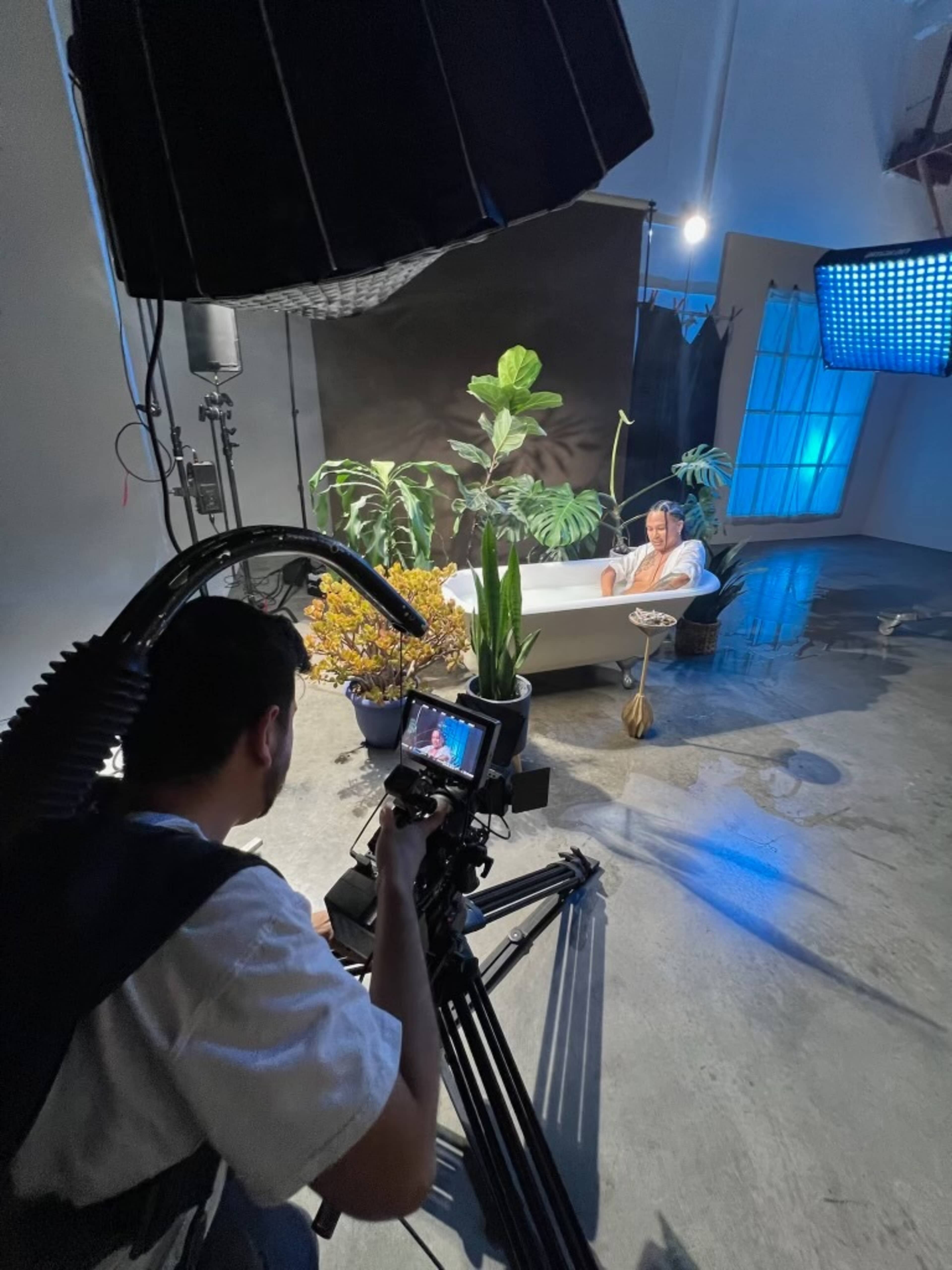 A person is sitting in a bathtub surrounded by plants while a cameraman records the scene in a studio setting.